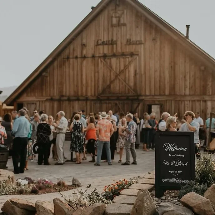 People gathered outside a large wooden barn for a wedding reception, with a welcome sign in the foreground.