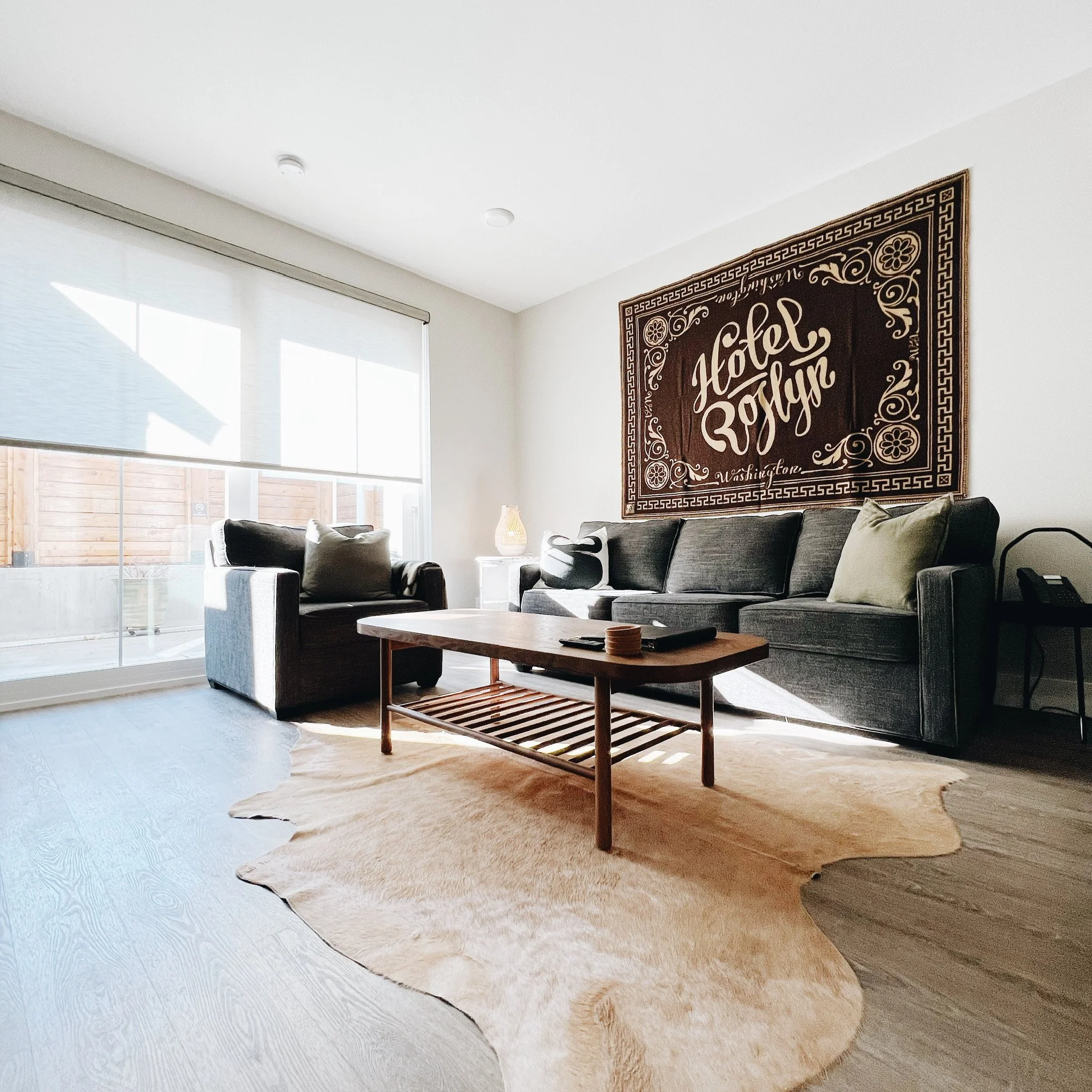 A King Patio room at Hotel Roslyn in Roslyn, WA. Living room with large window, beige rug, gray couch, wooden coffee table, and a decorative wall hanging.