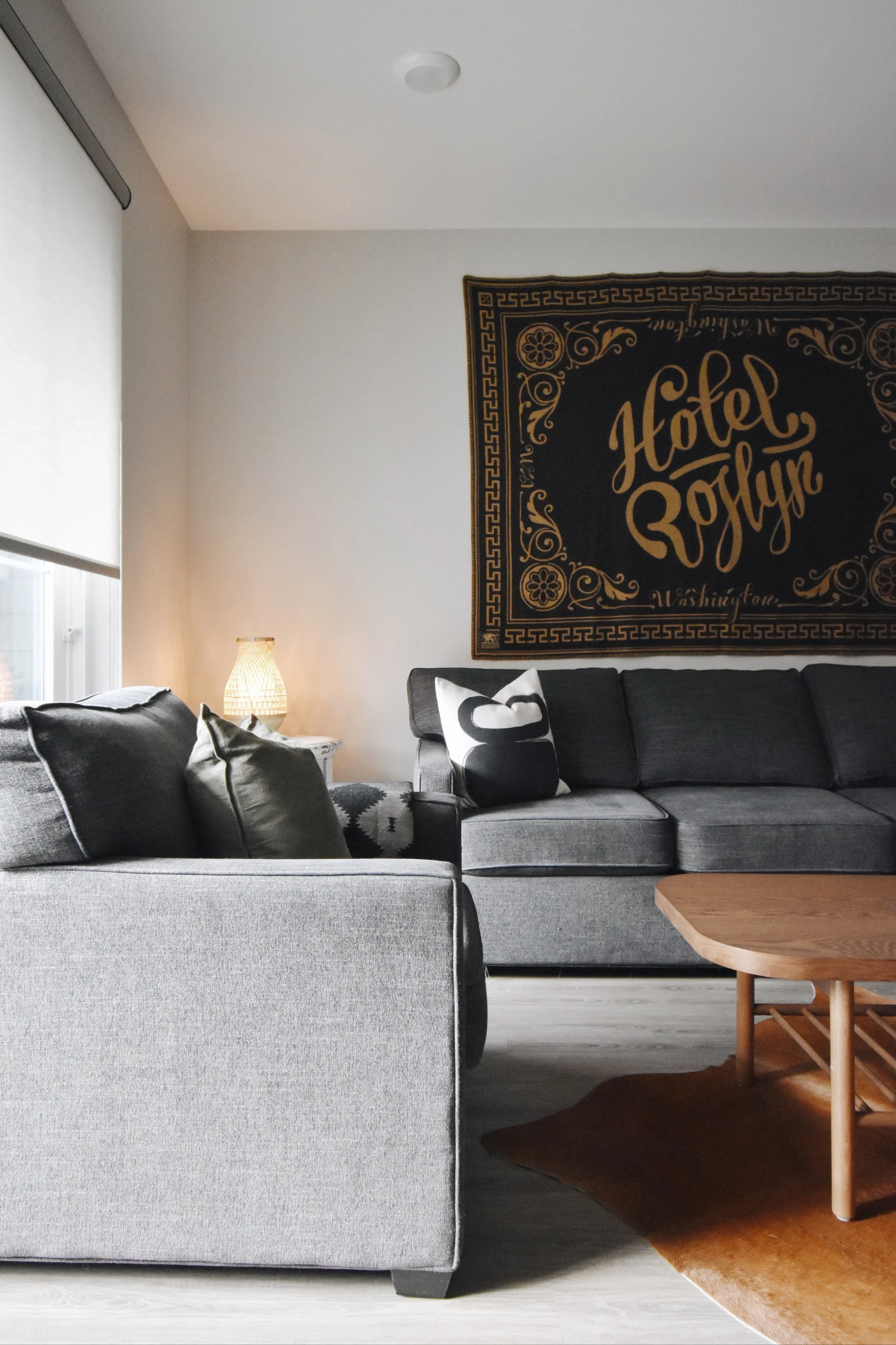 A living room with a gray sofa with pillows, a wooden coffee table, a wall hanging that reads "Hotel Roslyn" with decorative elements, a side table with a lamp, a window with a blind, a dark brown area rug, and a light-colored floor.