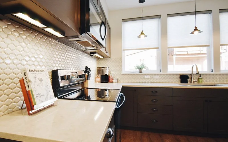 A King Patio room at Hotel Roslyn in Roslyn, WA. Kitchen with white hexagonal tile backsplash, dark lower cabinets, white countertops, and three windows with blinds and hanging pendant lights.