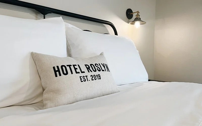 A close-up of a bed with white bedding and a decorative pillow that reads 'HOTEL ROSLYN EST. 2019'.