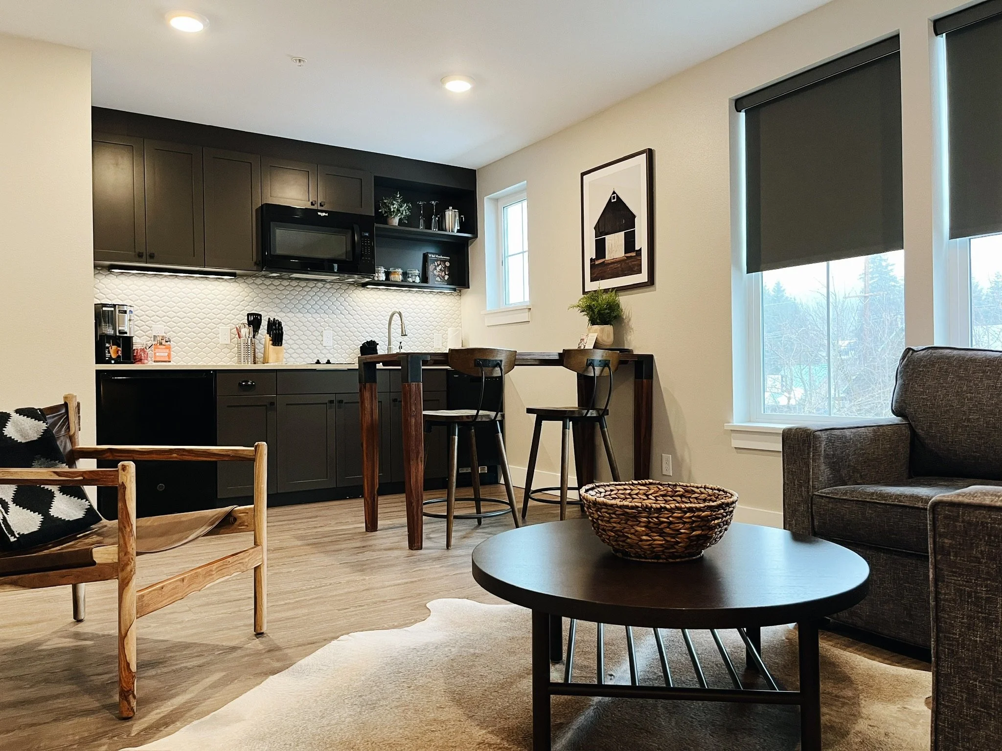 A 2nd Floor King Suite room at Hotel Roslyn, in Roslyn, WA. Interior of a modern apartment featuring a kitchen with dark cabinets, a white textured backsplash, and kitchen appliances, adjacent to a living area with a sofa, coffee table, and decor.