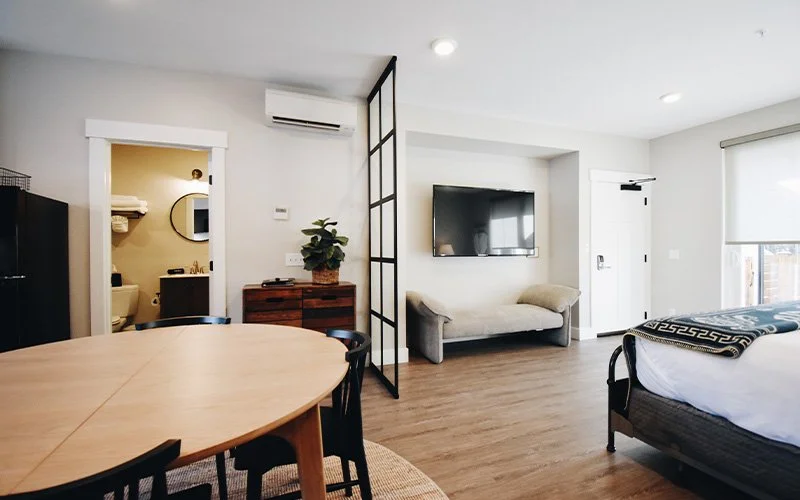 A King Patio room at Hotel Roslyn in Roslyn, WA. Modern living space with a dining table, a TV on the wall, a gray sofa, a bed with a patterned blanket, and a bathroom visible through an open doorway.