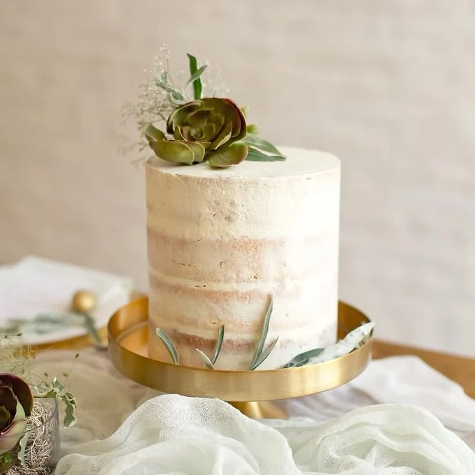 A minimalist nude-colored cake with thin layers and a succulent decoration on top, placed on a gold cake stand with green leaves and fabric around it.