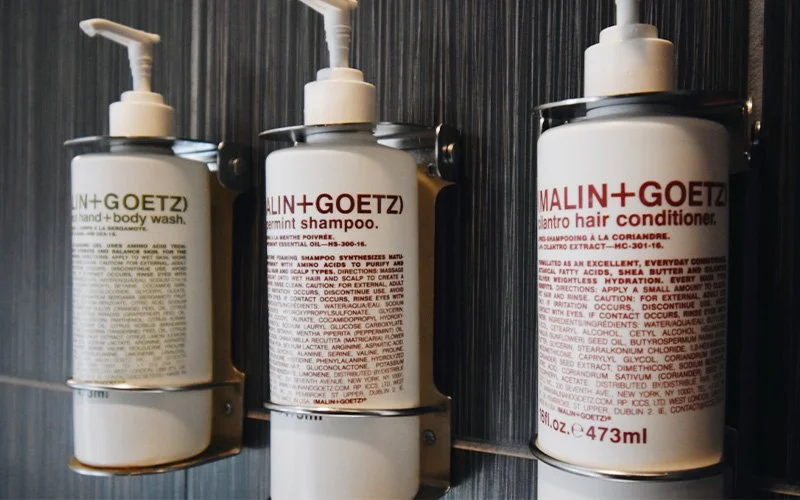 Three white bottles of Malin + Goetz hair care products mounted on a black wall shelf, including scalp shampoo, ginger shampoo, and cilantro hair conditioner, each with pump dispensers.