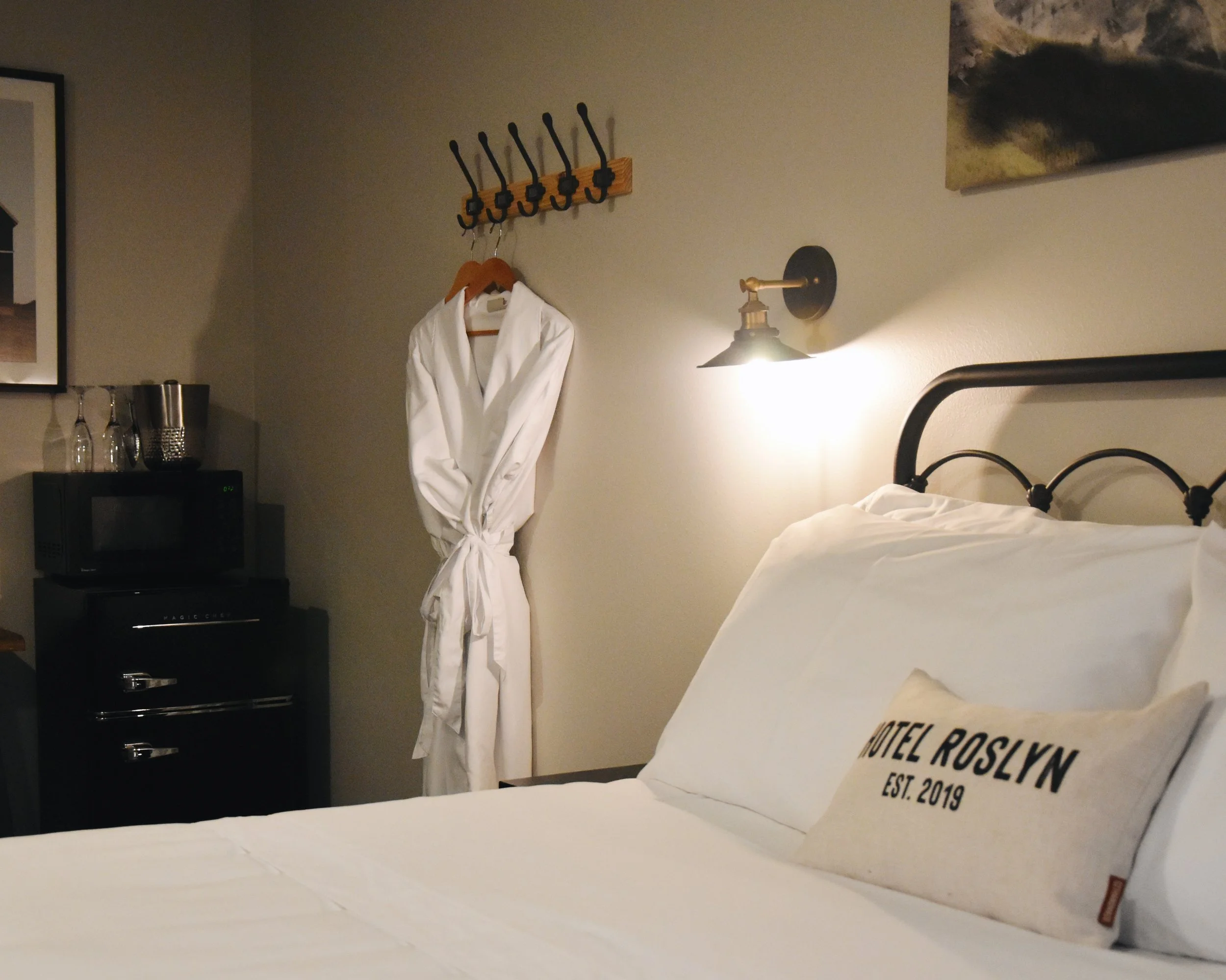 A hotel room with a bed, a white bathrobe hanging on a hook on the wall, a microwave on a black dresser, glasses and a metal container on top of the dresser, a wall-mounted light fixture, part of a painting on the wall, and a pillow with text reading 'Hotel Roslyn Est. 2019'.