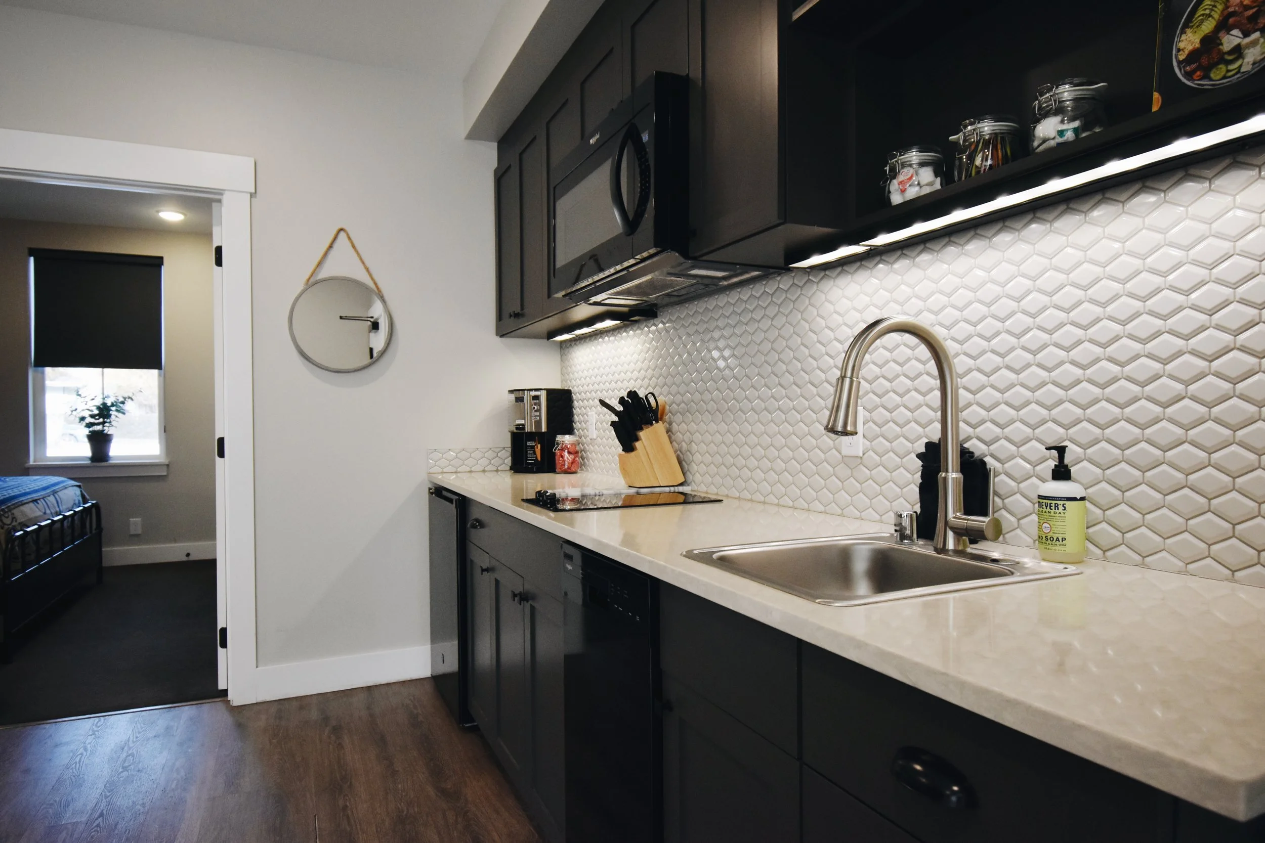 Modern kitchen with dark cabinets, white honeycomb tile backsplash, stainless steel sink, black marble countertop, and various kitchen items including a knife block, soap dispenser, and jarred items. A window and a doorway are visible in the background.