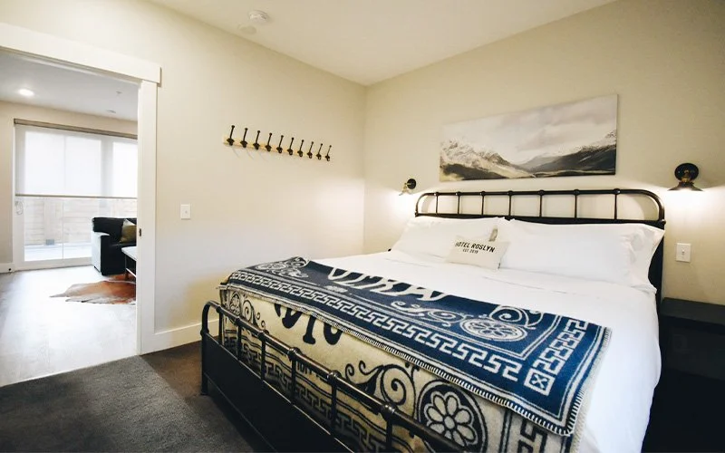 A King Patio room at Hotel Roslyn in Roslyn, WA. Bedroom with a black metal bed frame, white bedding, a decorative bed runner, wall-mounted lamps, a large landscape painting, and a view into a living room with a sliding glass door and sofa.