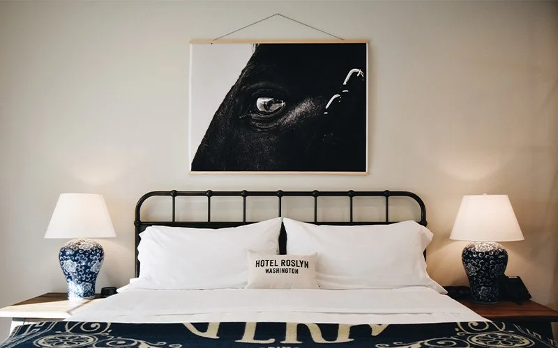 A King Patio room at Hotel Roslyn in Roslyn, WA. A hotel room featuring a black iron bed with white bedding and pillows, two blue and white lamps on wooden nightstands, a framed artwork of a horse's eye hanging above the bed, and a pillow.