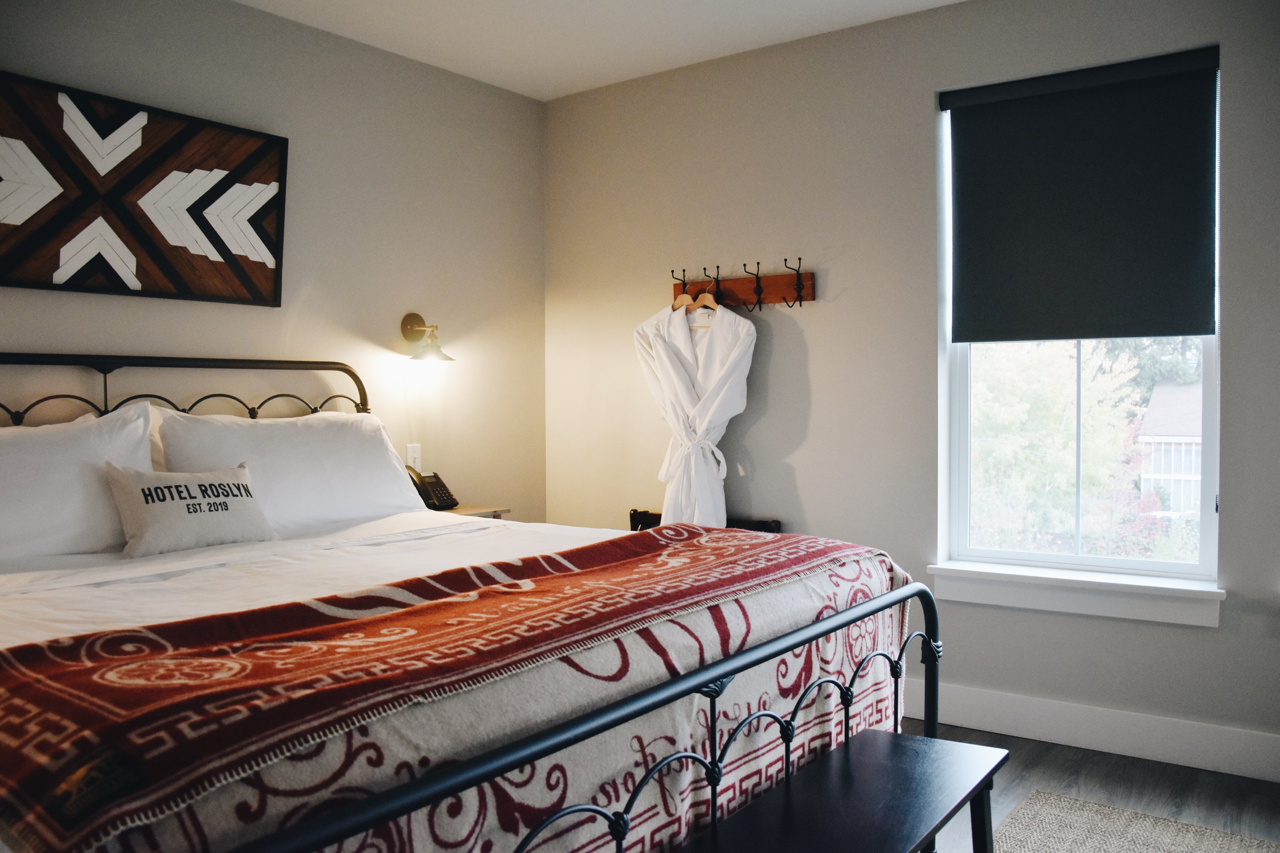 A hotel room with a bed featuring white bedding and pillows, a decorative wall art, a side table with a phone, a wall-mounted lamp, and a window with a closed dark curtain.