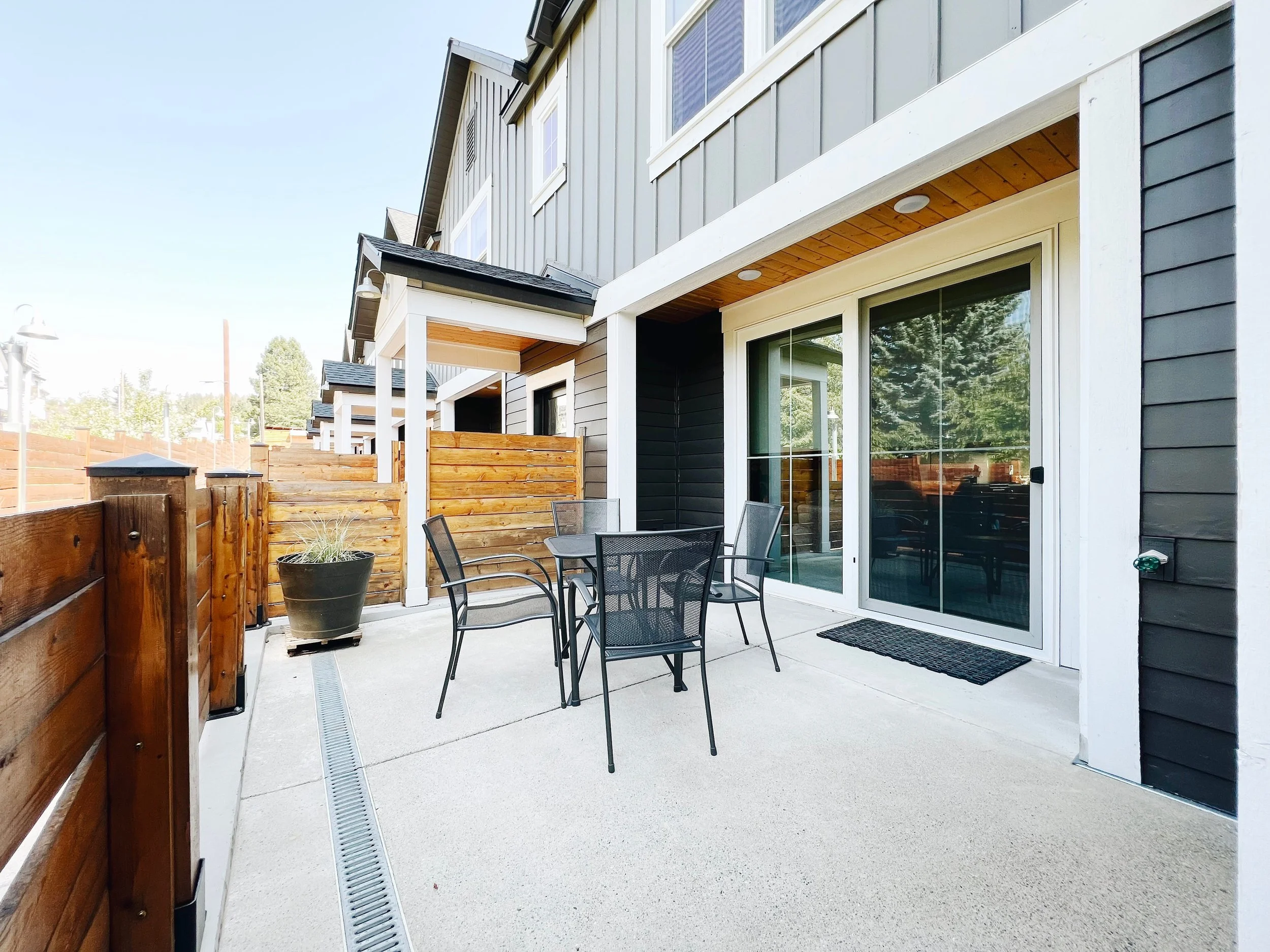A patio area with a table and chairs in front of a building with gray siding and a wooden fence.