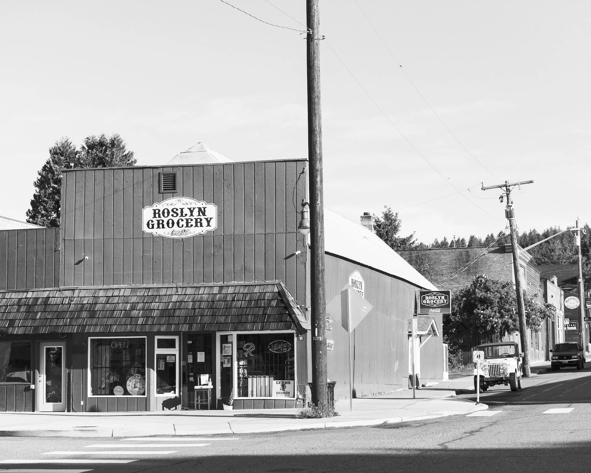 Black and white photo of a small grocery store called Roslyn Grocery. The storefront has large windows and a door, with signs indicating it is open. There is a sidewalk in front, with a curb, and a street with a car parked along the curb. Utility poles and power lines are visible in the scene, along with a tree and rooftops of nearby buildings under a clear sky.
