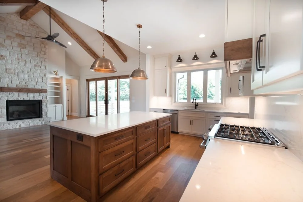 Kitchen with a stained wood island and copper pendant lights, open to a living room with a stone fireplace and exposed ceiling beams