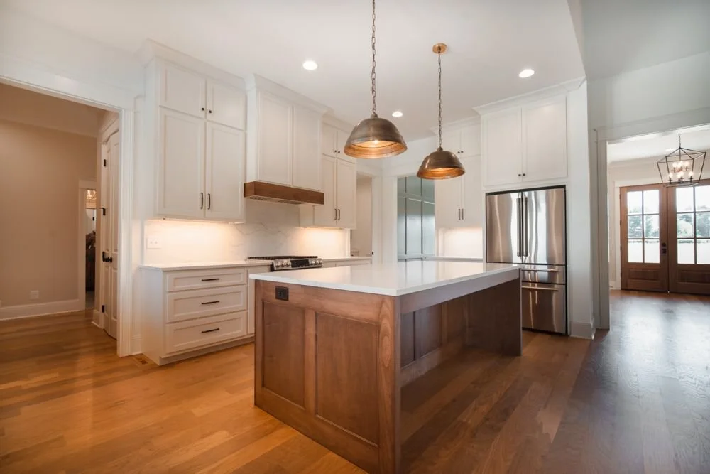 Stained wood kitchen island under two copper pendants with white cabinets, stainless refrigerator and a wood range hood