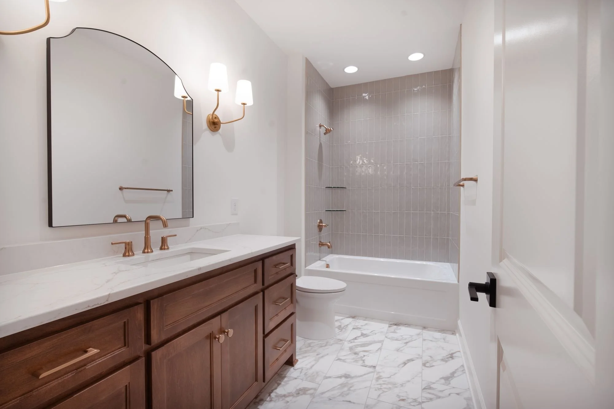 Bathroom with a wood vanity, arched mirror, brass fixtures, marble-look floor and a tiled tub and shower