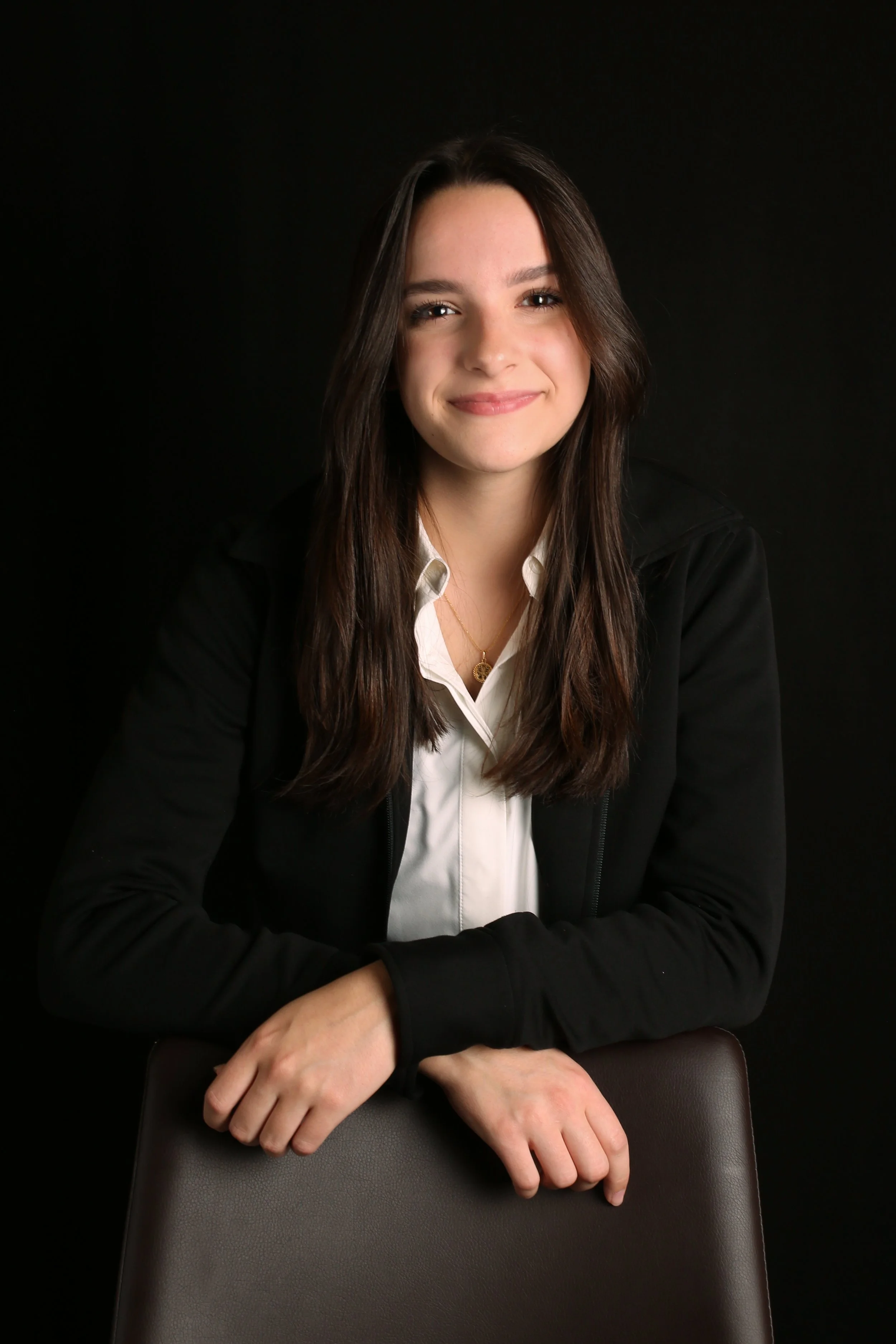 A young woman with long brown hair, smiling softly, sitting in front of a black background, wearing a black blazer and a white blouse.