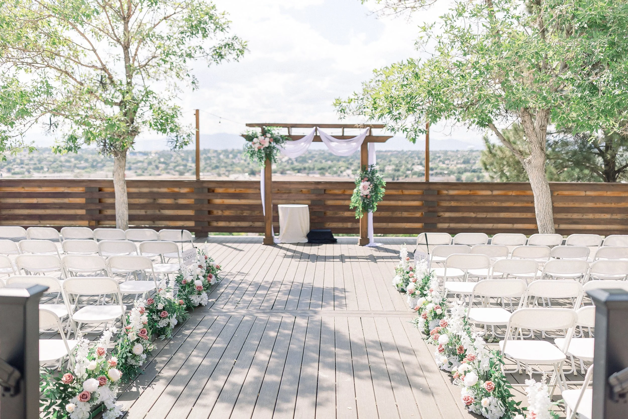 An outdoor wedding ceremony setup on a wooden deck framed by two trees with lush green leaves. The altar is decorated with white fabric and floral arrangements, with chairs arranged in rows for guests. There are floral decorations along the aisle. Th