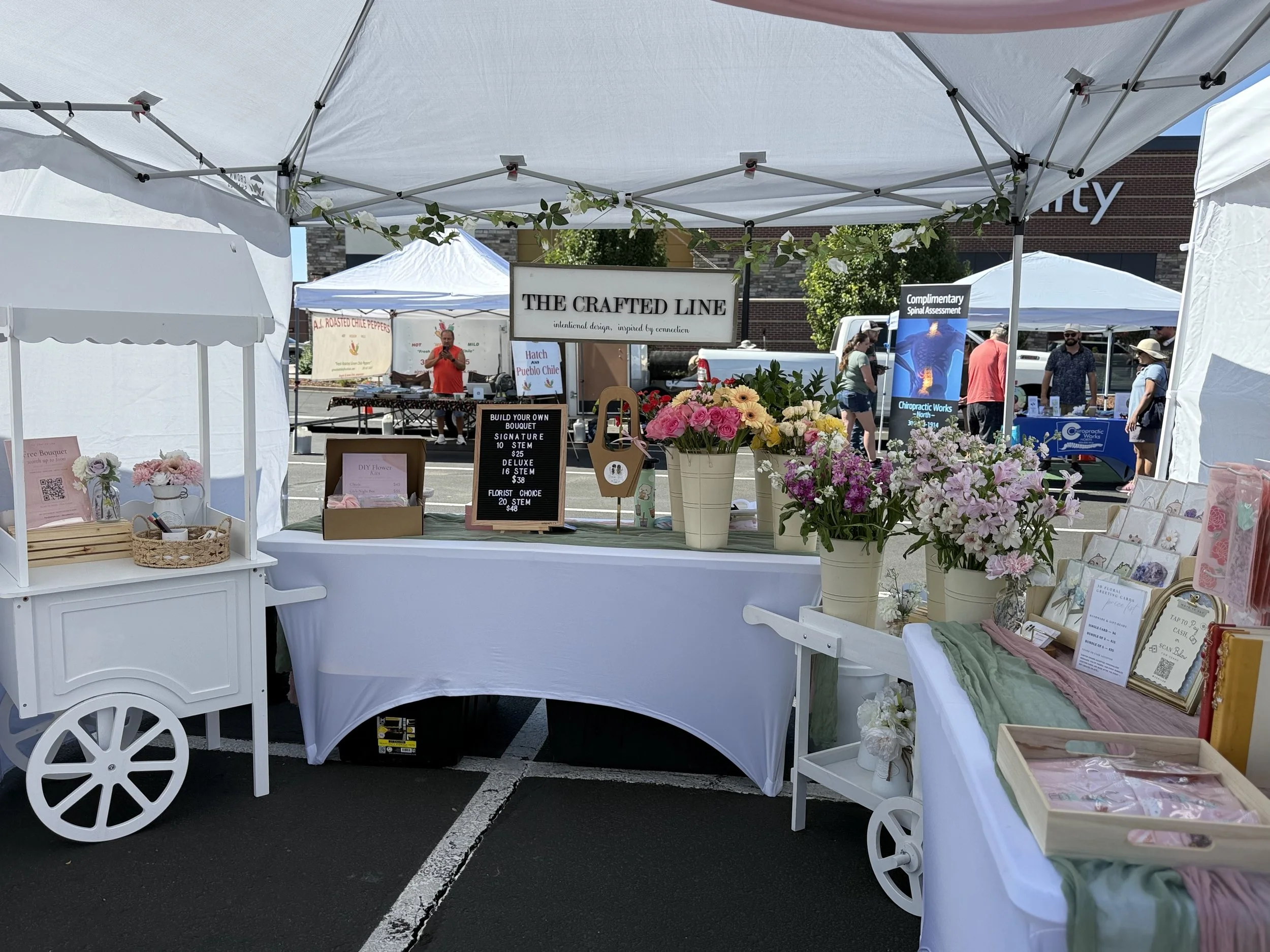 A full view of your market booth featuring florals, framed signage, and curated décor under a white canopy — highlighting your signature handcrafted, boutique vendor style.