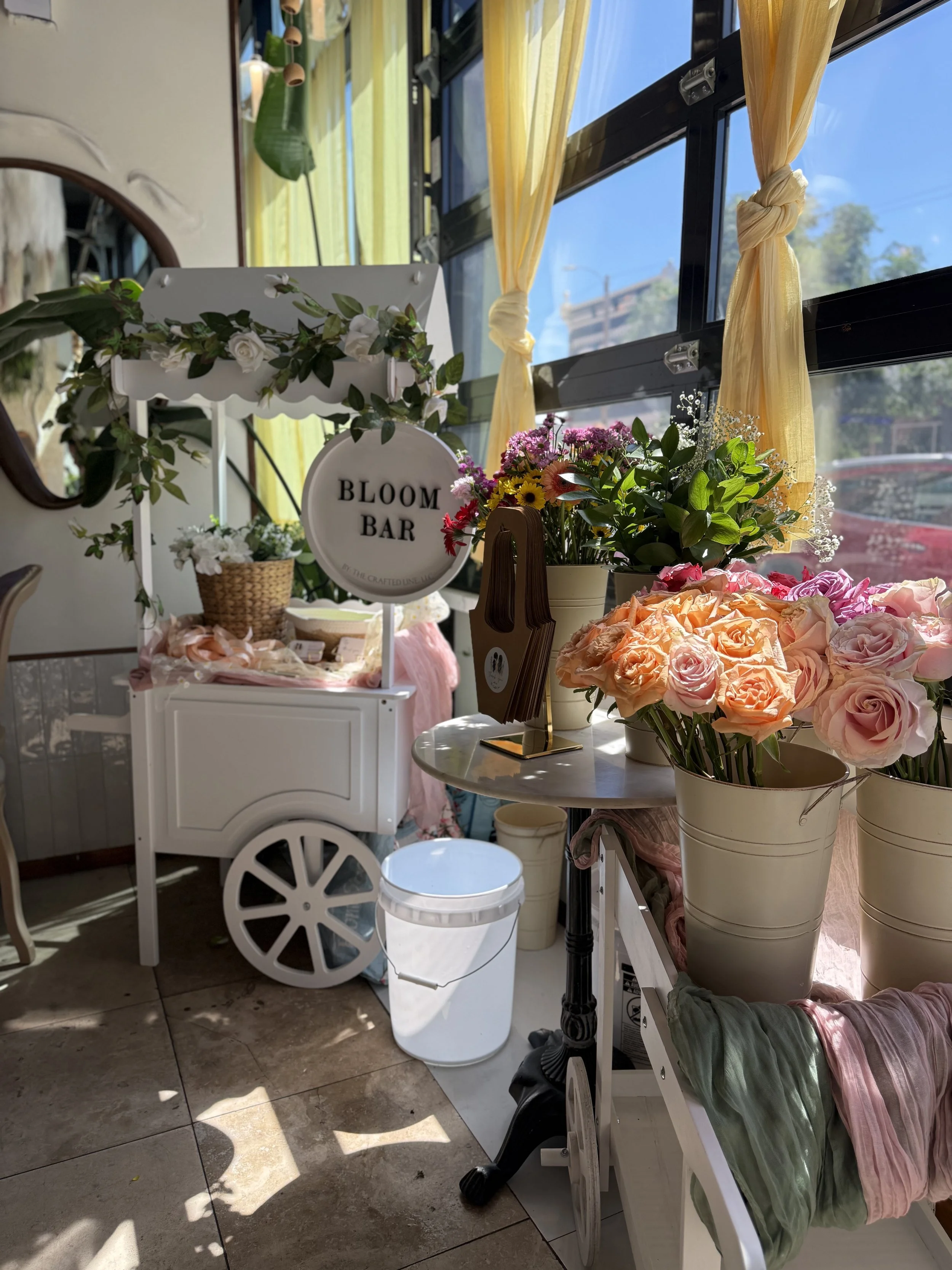 The full bloom bar setup — a white cart topped with a "Bloom Bar" sign, flanked by galvanized buckets overflowing with peonies, ranunculus, and mixed blooms in coral, pink, and cream. Gold streamer backdrop and flower wall behind.