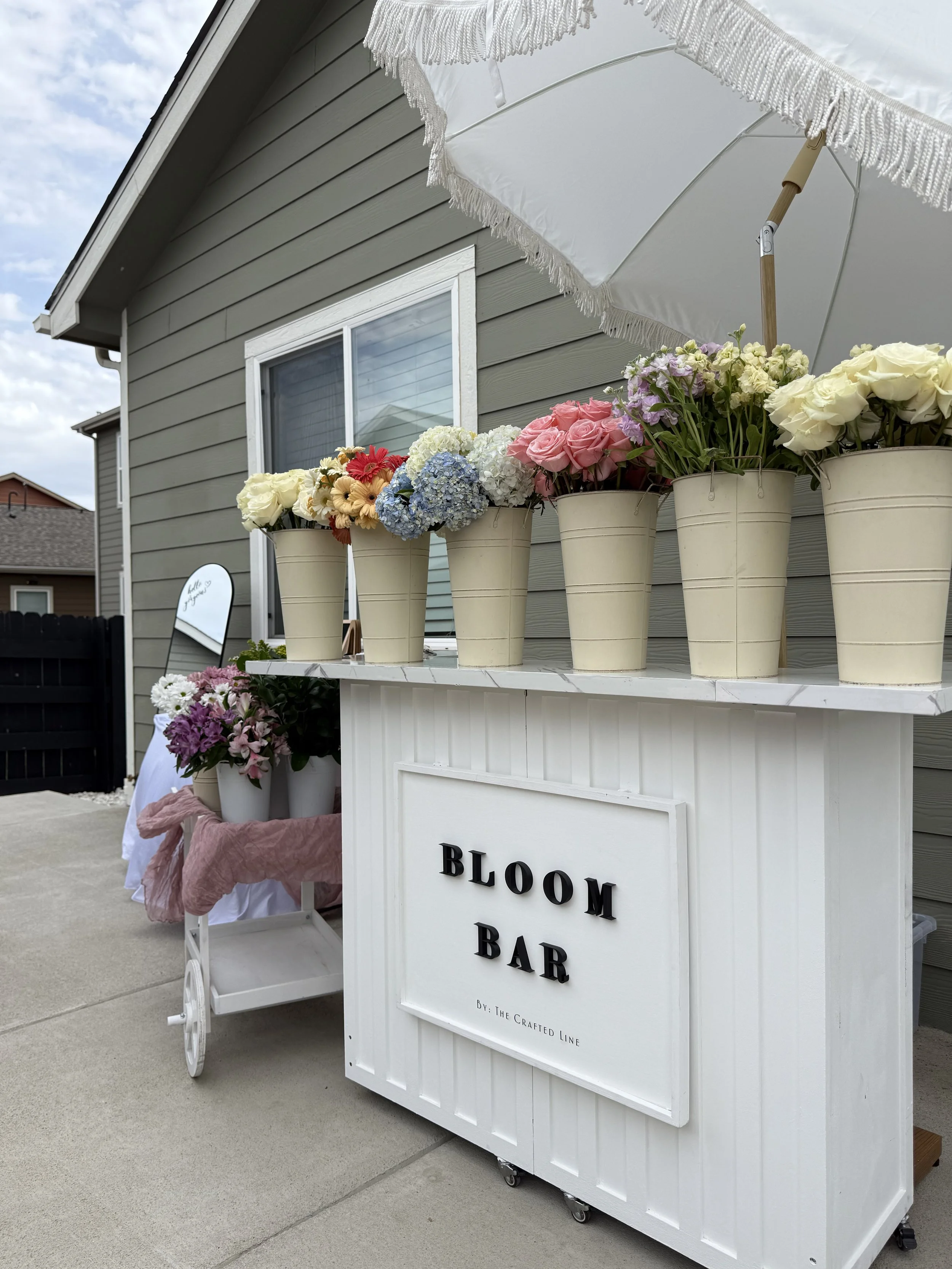 A white mobile flower cart with a sign that reads 'Bloom Bar' by The Crafted Line, decorated with various bouquets of flowers, including roses, hydrangeas, and other blooms, set outside against a gray house.