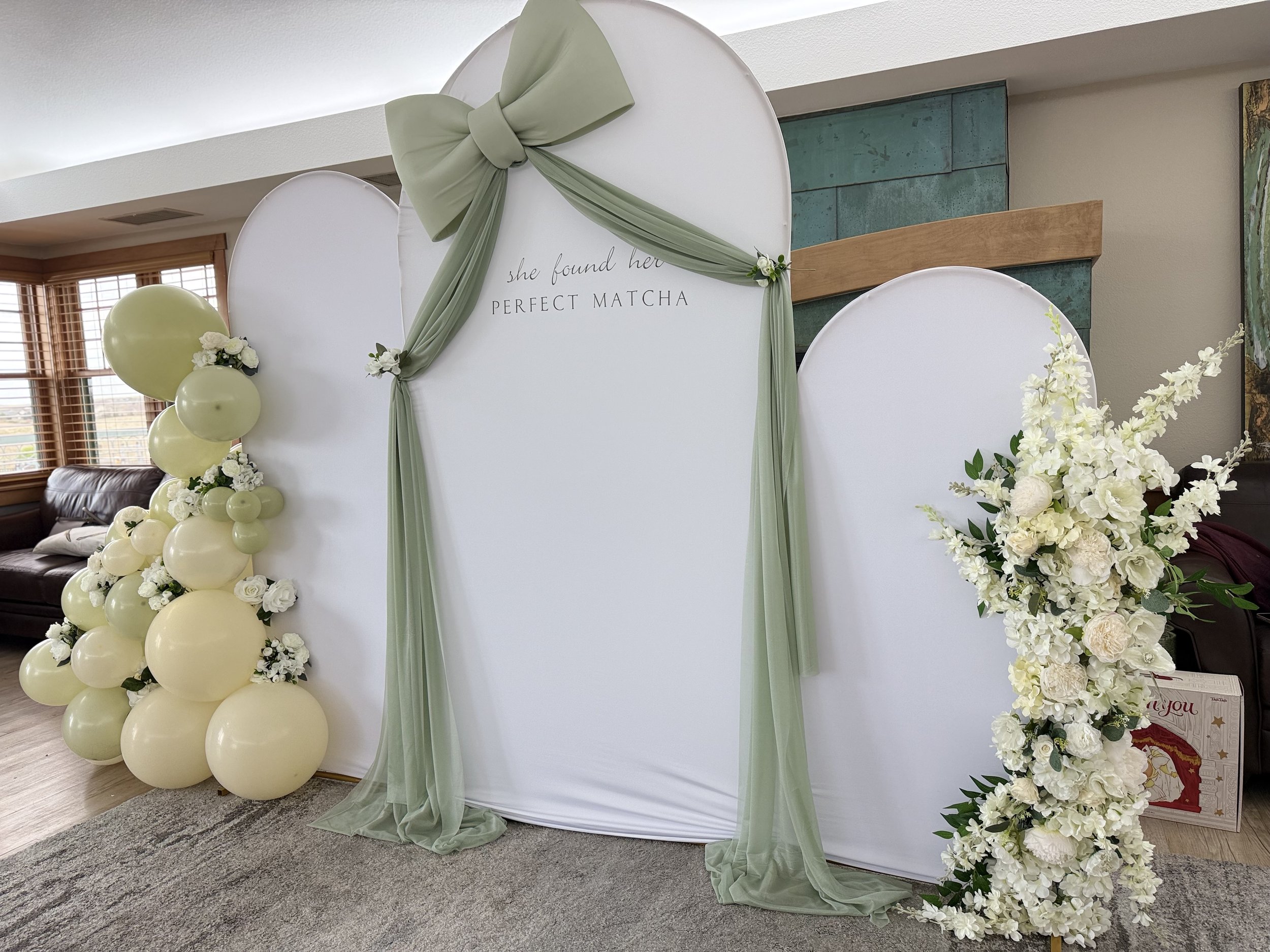 A wedding backdrop decorated with green and white balloons, flowers, and draped fabric, with a sign that reads 'she found her perfect matcha.'