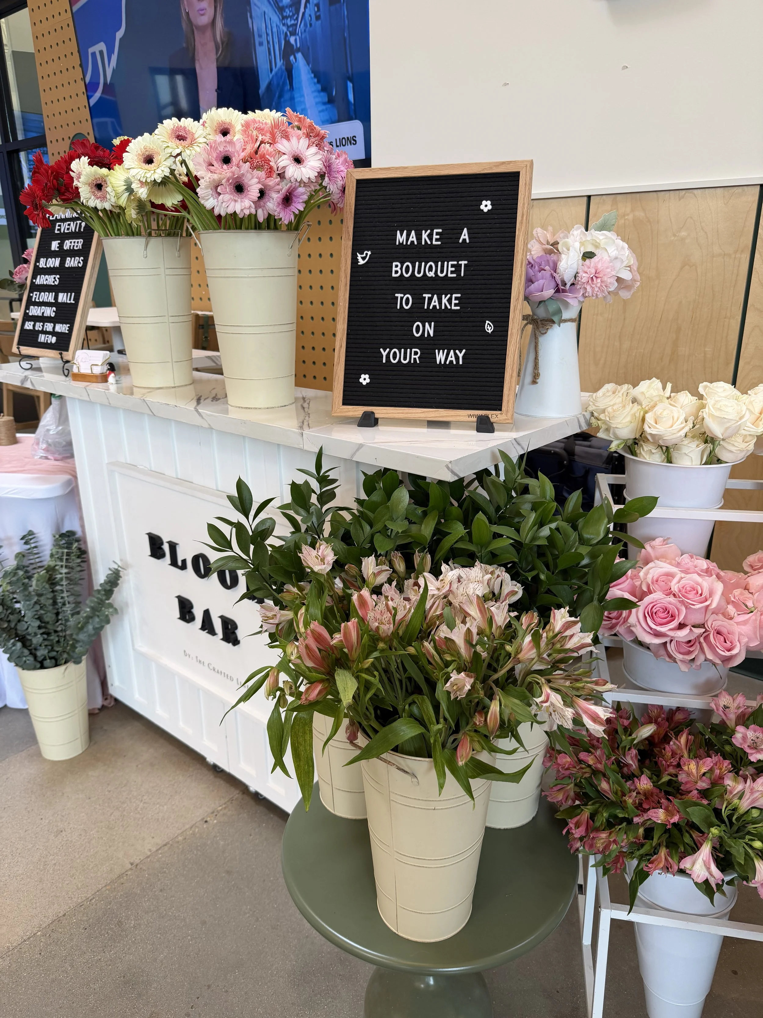 Various flower arrangements in white pots on display at a floral shop, featuring pink, white, and purple flowers, with a sign reading 'Make a bouquet to take on your way' and some greenery.