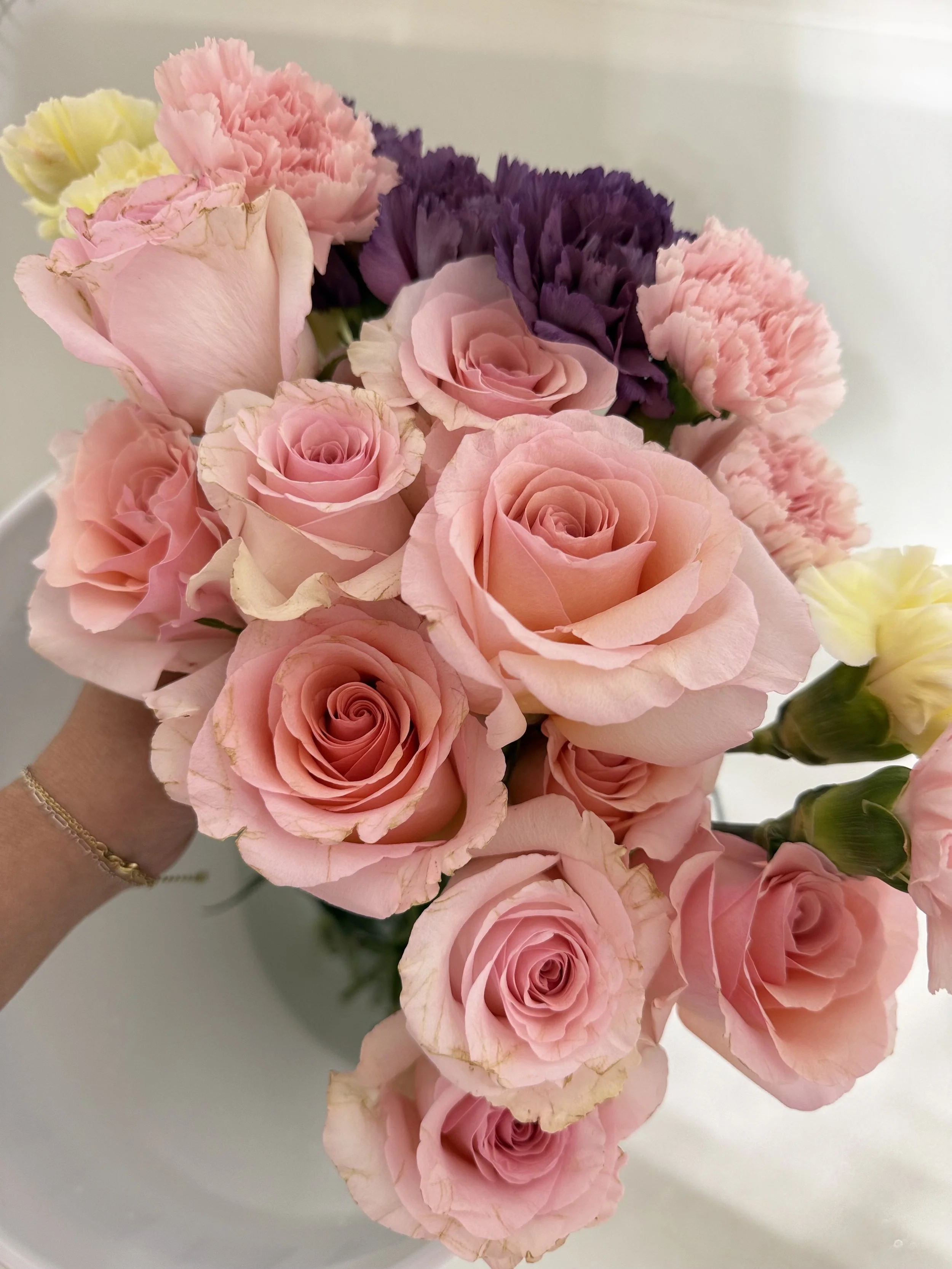 A bouquet of pink roses, purple carnations, pink carnations, and a pale yellow flower against a light background.