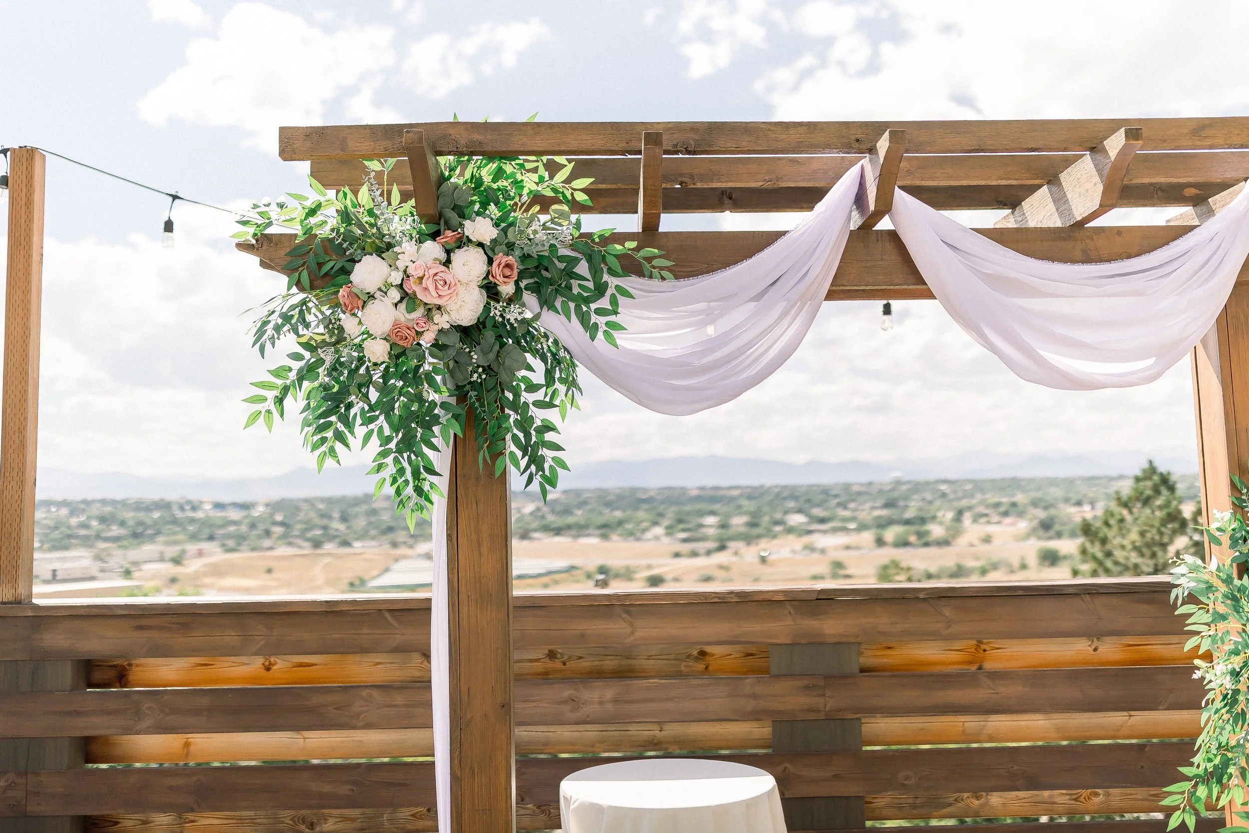 Wedding arch decorated with pink and white flowers and green foliage, draped with white fabric, overlooking a landscape with cloudy sky, distant hills, and buildings.