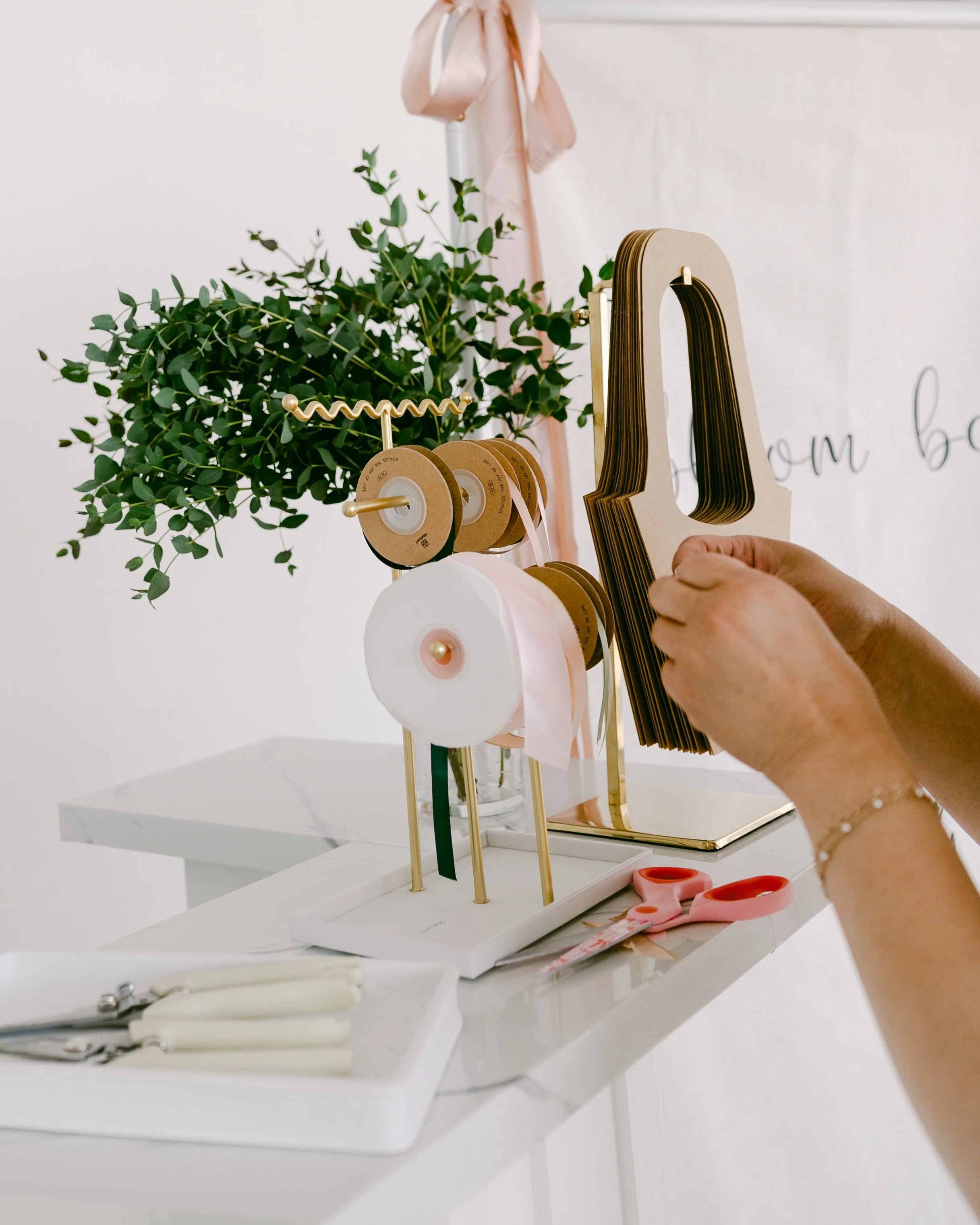A soft, intimate behind‑the‑scenes moment showing hands working with ribbon spools and floral materials on a bright white table. The greenery in the background adds a fresh, organic feel, highlighting the craftsmanship that goes into every bloom bar 