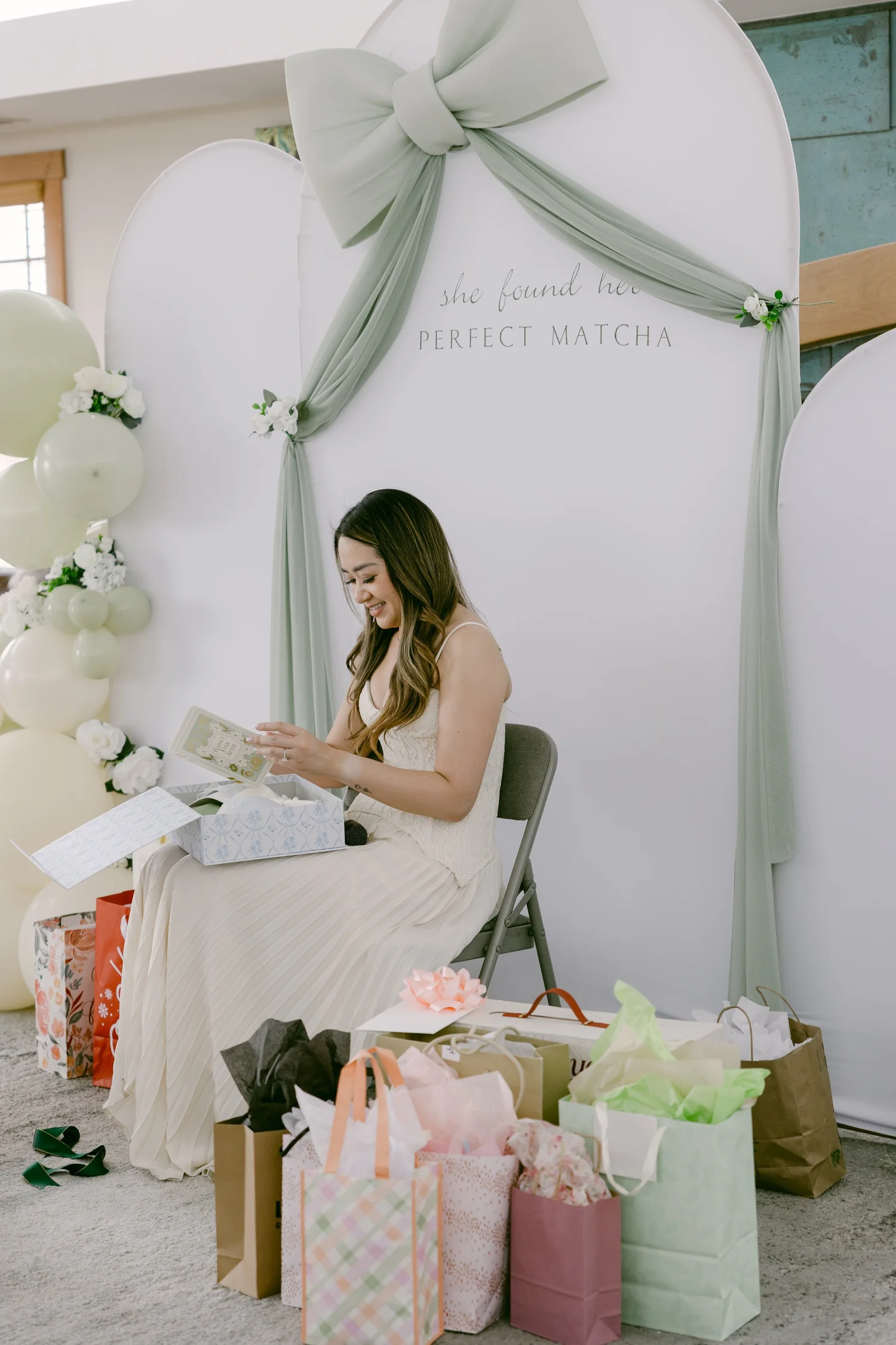 Woman in cream dress at a celebration with gift bags, a decorated backdrop with balloons and drapery