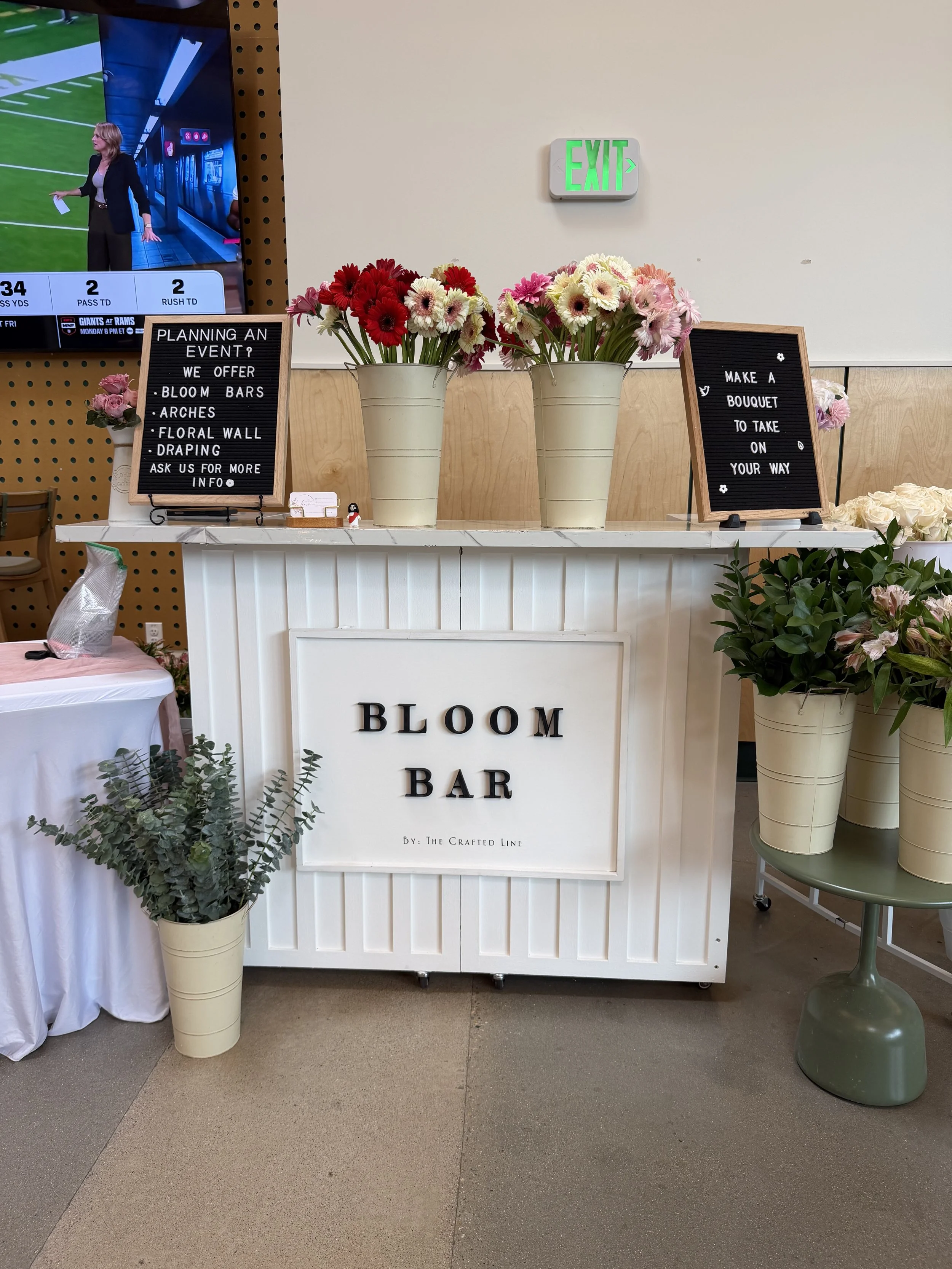 A floral display at a wedding or event reception with a white table and large cream-colored flower pots holding red, pink, and white flowers. Two chalkboards with white lettering advertise event planning services and bouquet preparation. A green side