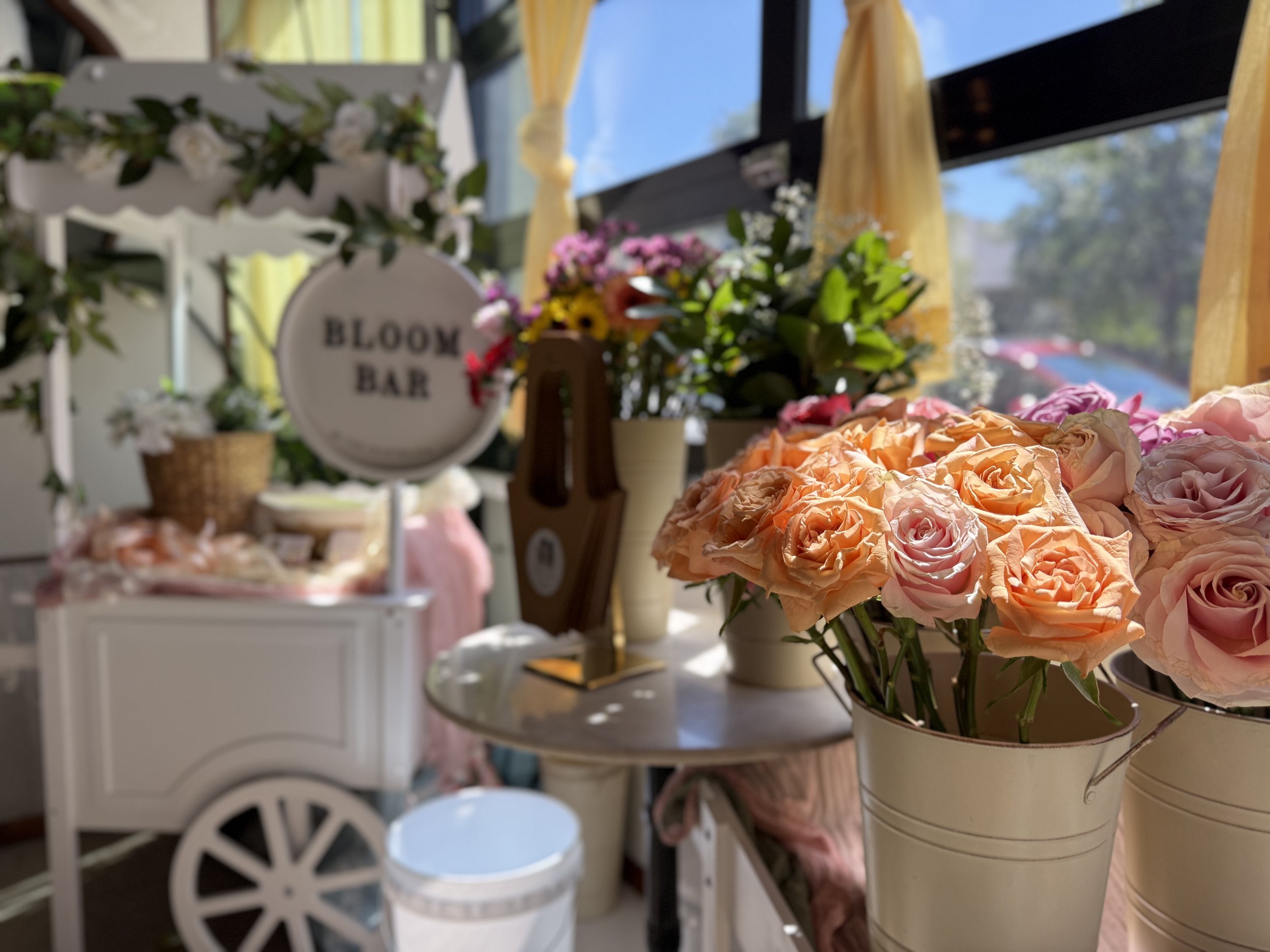 A styled detail shot of the "Bloom Bar" the white cart and signage softly blurred in the background, with a galvanized bucket of coral garden roses and mixed stems in sharp focus in the foreground. Flower wall and gold streamers frame the scene.