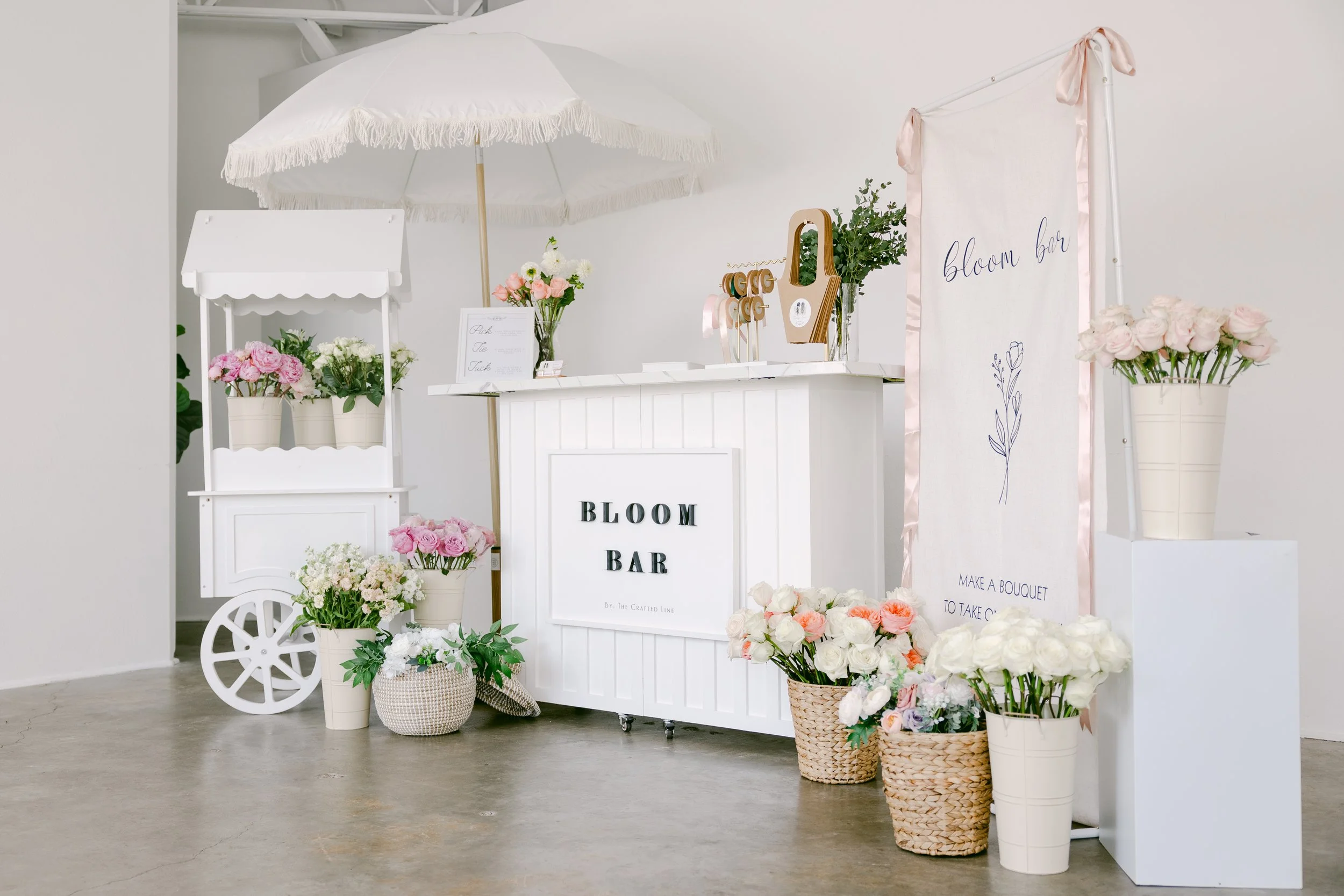 A beautifully styled white bloom bar setup featuring a crisp “BLOOM BAR” sign, soft florals, and a chic white umbrella. The overall look is clean, elegant, and inviting — the perfect modern floral experience for events.