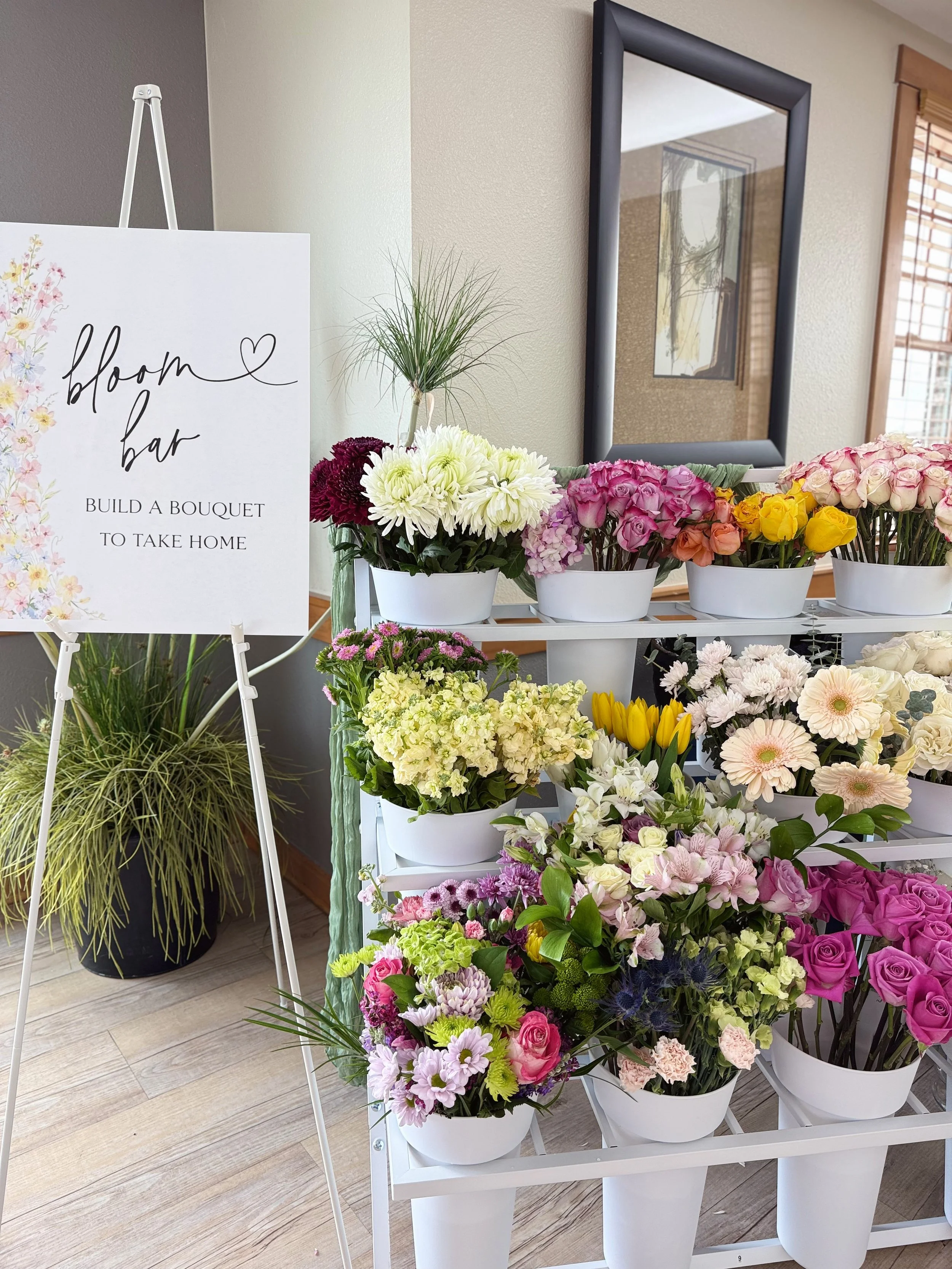 A display of various colorful flowers arranged in white pots and a white flower stand, with a sign reading 'bloom bar' and 'build a bouquet to take home' near a mirror and window with wooden blinds, in a bright room.