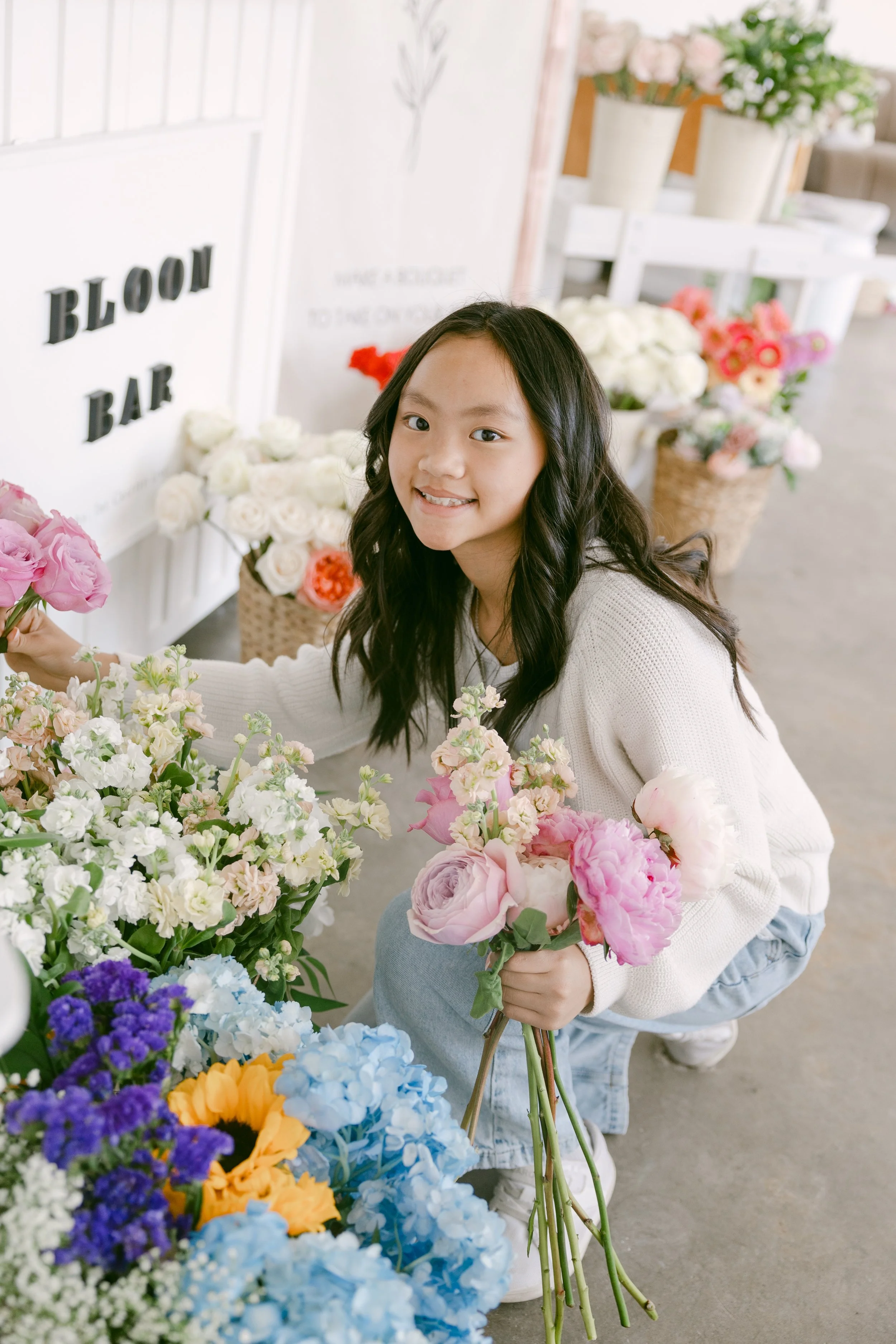 A candid, cozy shot of someone kneeling beside a floral display, holding a freshly made bouquet. The “BLOOM BAR” sign behind them ties the scene together, capturing the joy and creativity of choosing and arranging blooms.