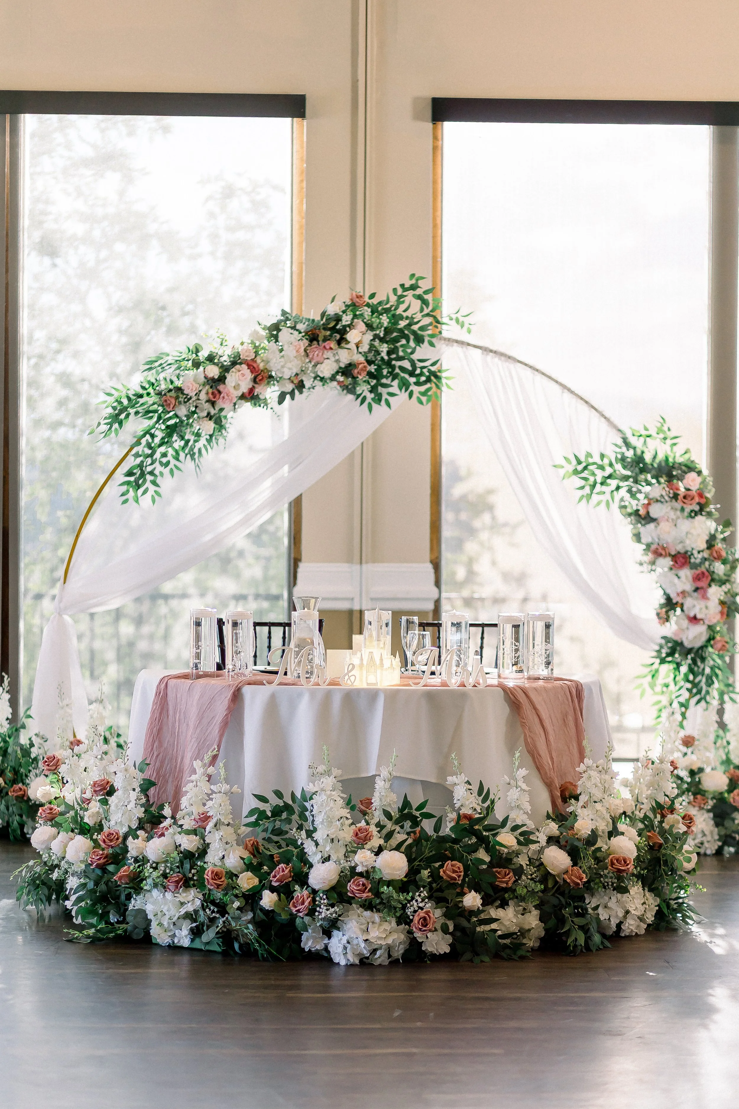 Wedding reception table decorated with pink and white flowers, greenery, and a draped pink-colored cloth, with a floral arch and windows in the background.