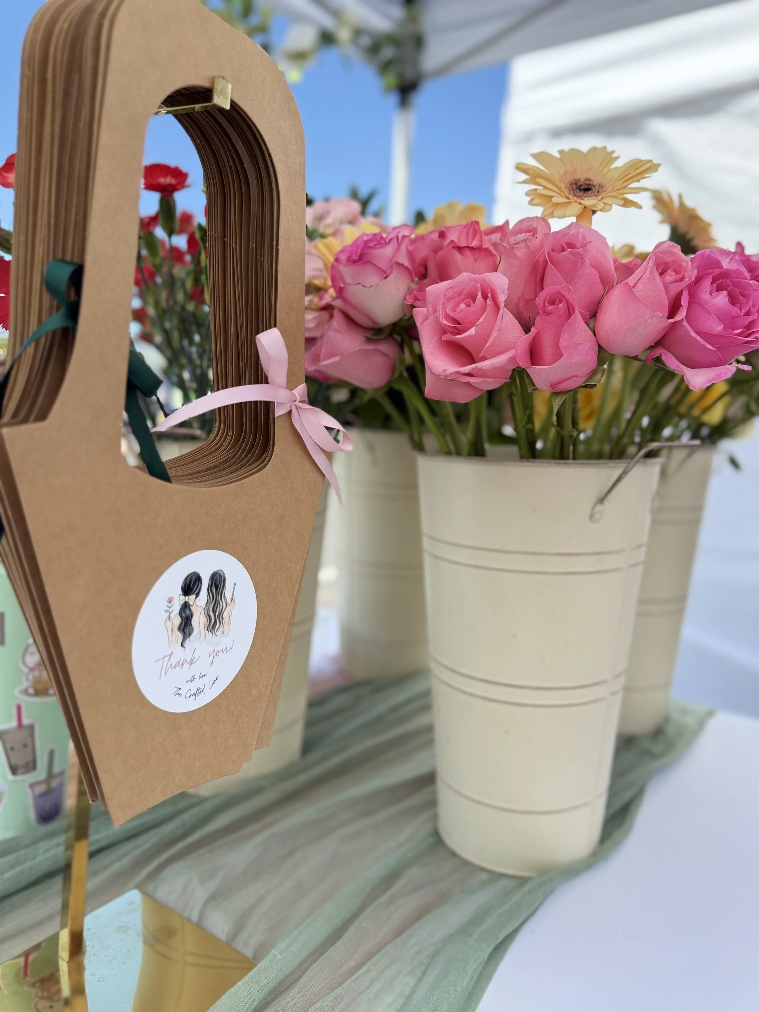 Blush and pink roses styled in cream buckets with a bee‑themed paper bag — an inviting detail shot from your floral bar setup.