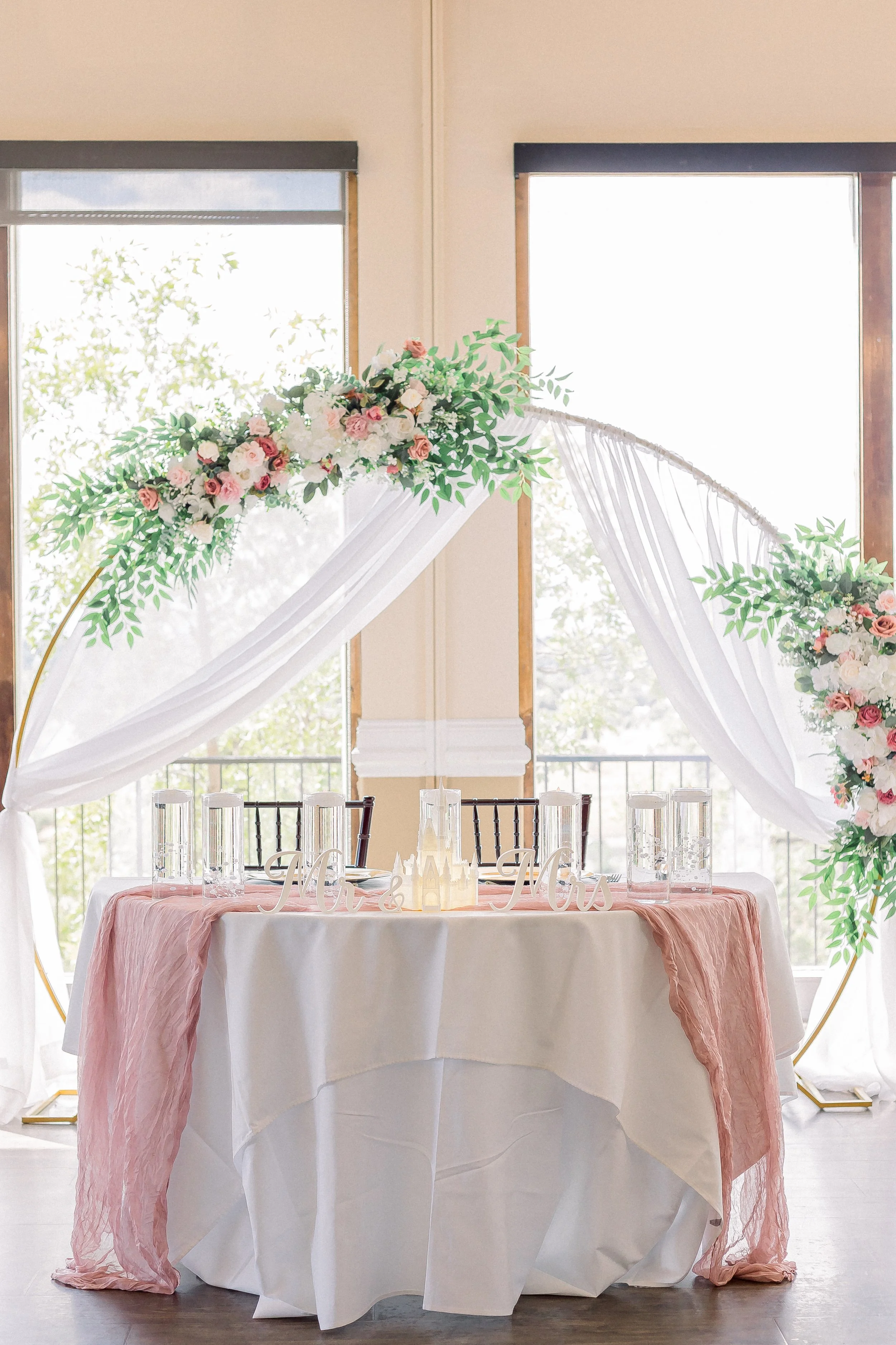 A wedding reception table decorated with white and pink flowers, white drapes, and a pink table runner, set in front of large windows with natural light.