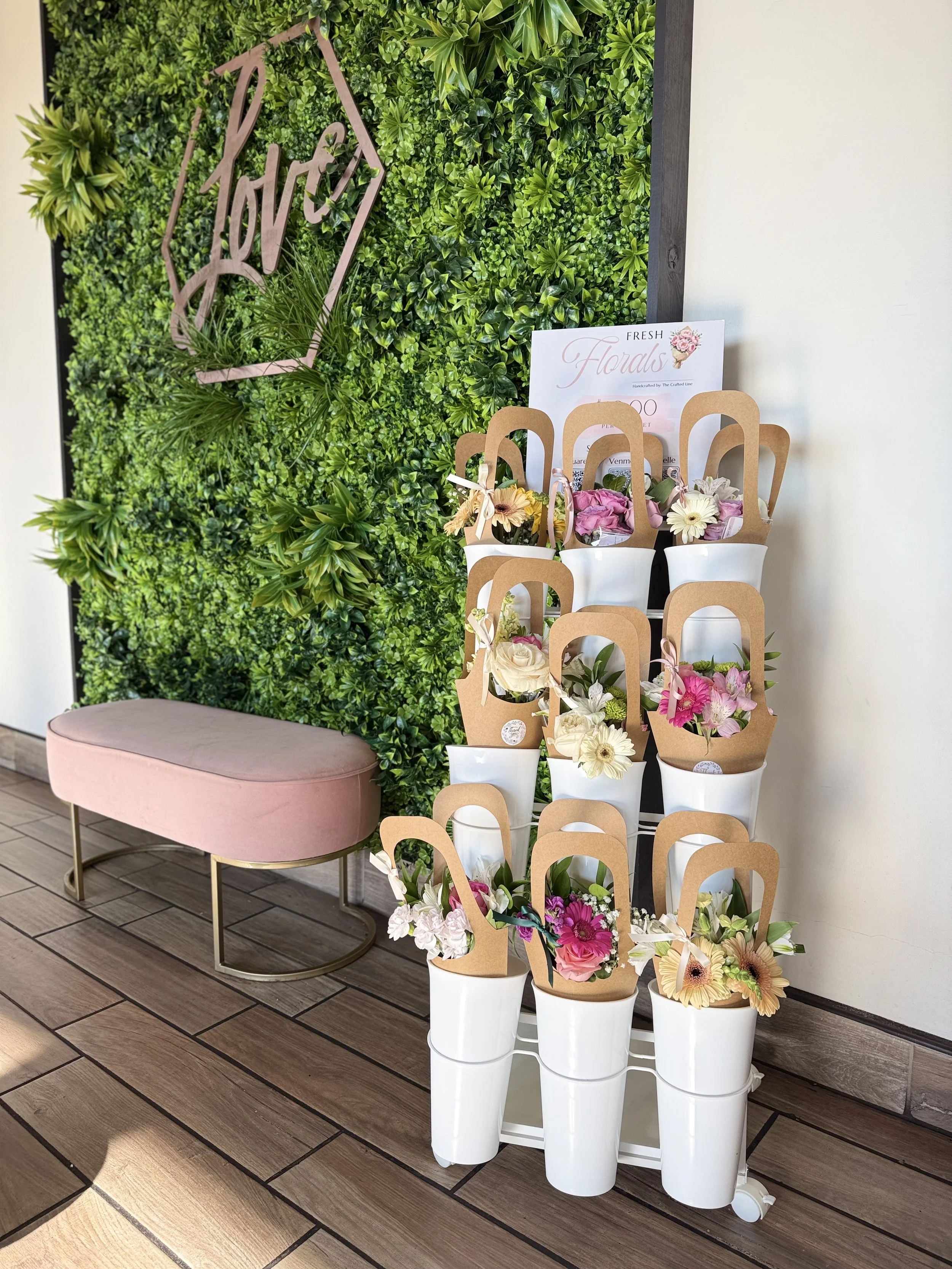 A soft, boutique‑style floral display featuring tiered white cylinders filled with brown‑paper‑wrapped mini bouquets, set against a lush greenery wall with a cursive “Love” sign and paired with a blush bench for a clean, romantic aesthetic.