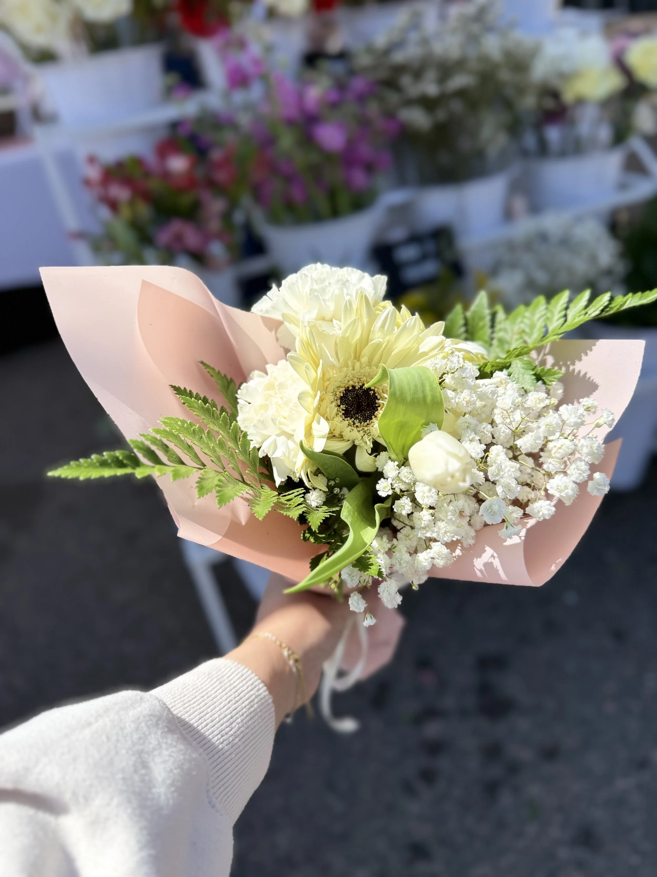 A person holding a bouquet of white and cream flowers wrapped in pink paper, outdoors, with blurred background of market stalls and colorful flowers.