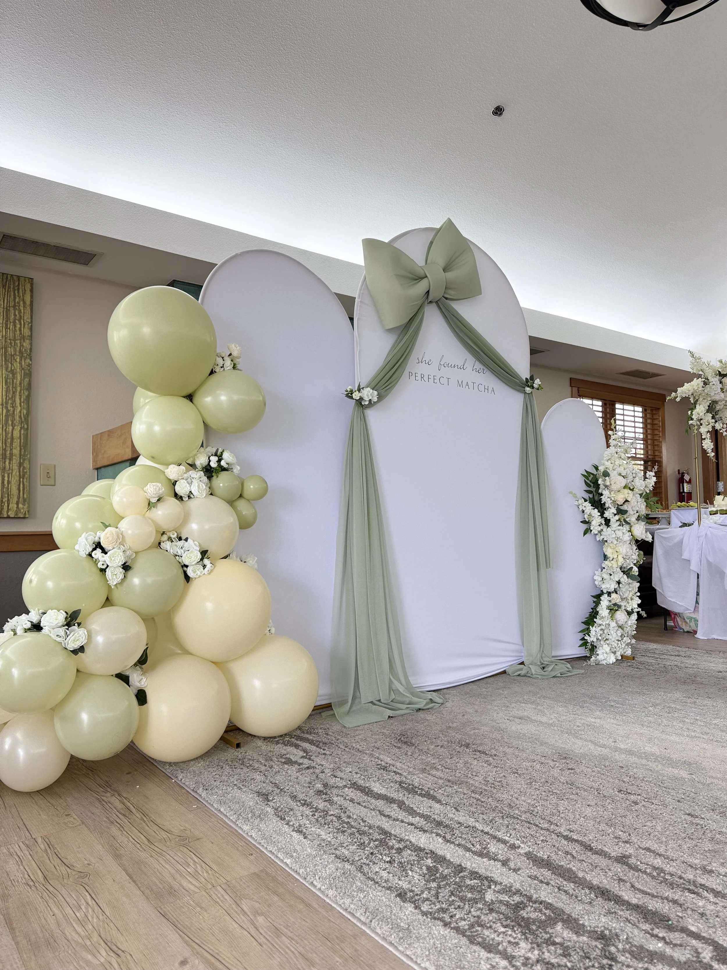 Decorative event backdrop with balloons, flowers, and draped fabric, featuring a bow and a sign that reads "she found her perfect match."