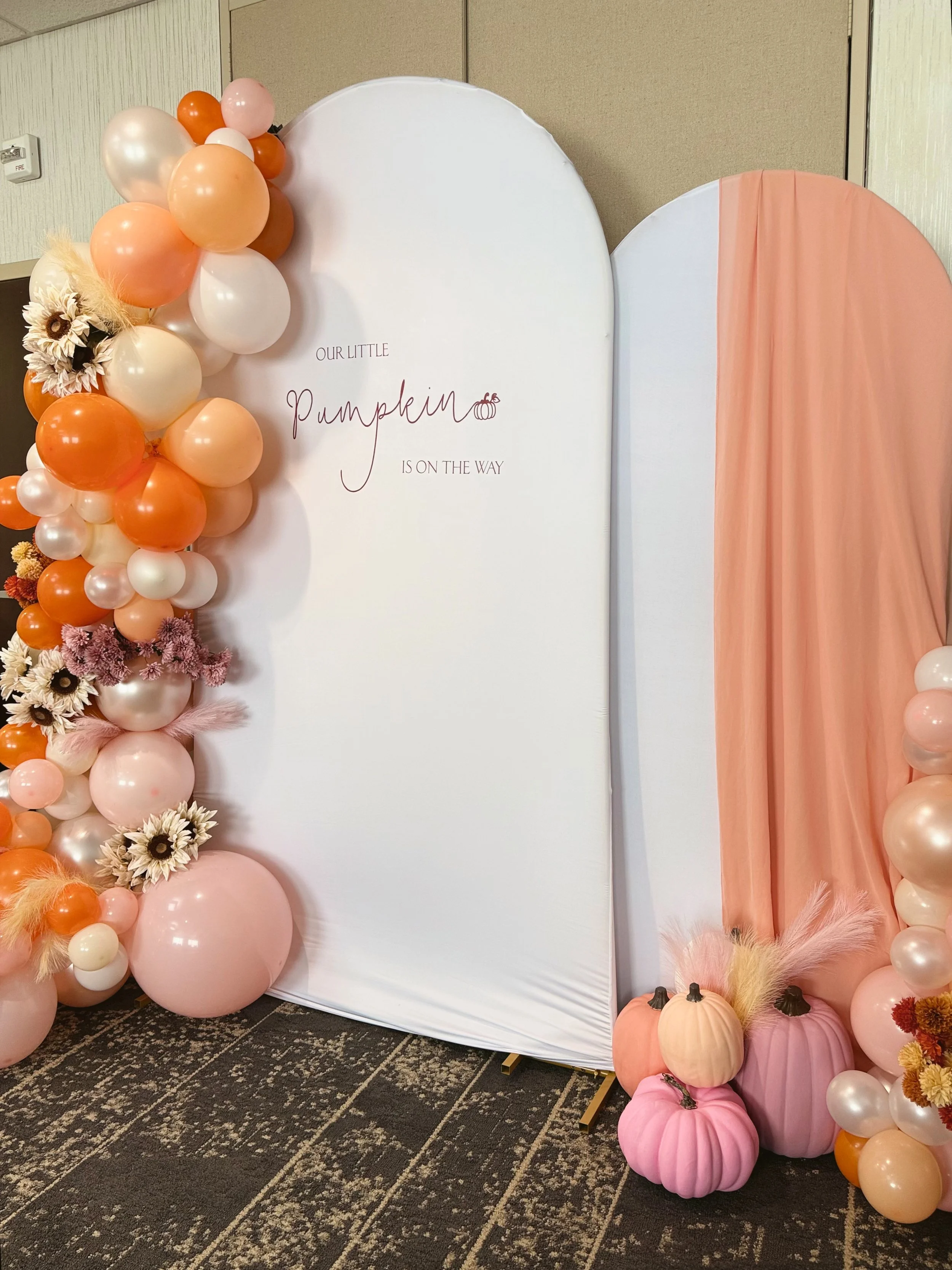 Decorative display featuring a balloon garland with peach, pink, white, and orange balloons, flowers, pumpkins, and fall foliage around a white backdrop with the text 'Our Little Pumpkin is on the Way'.