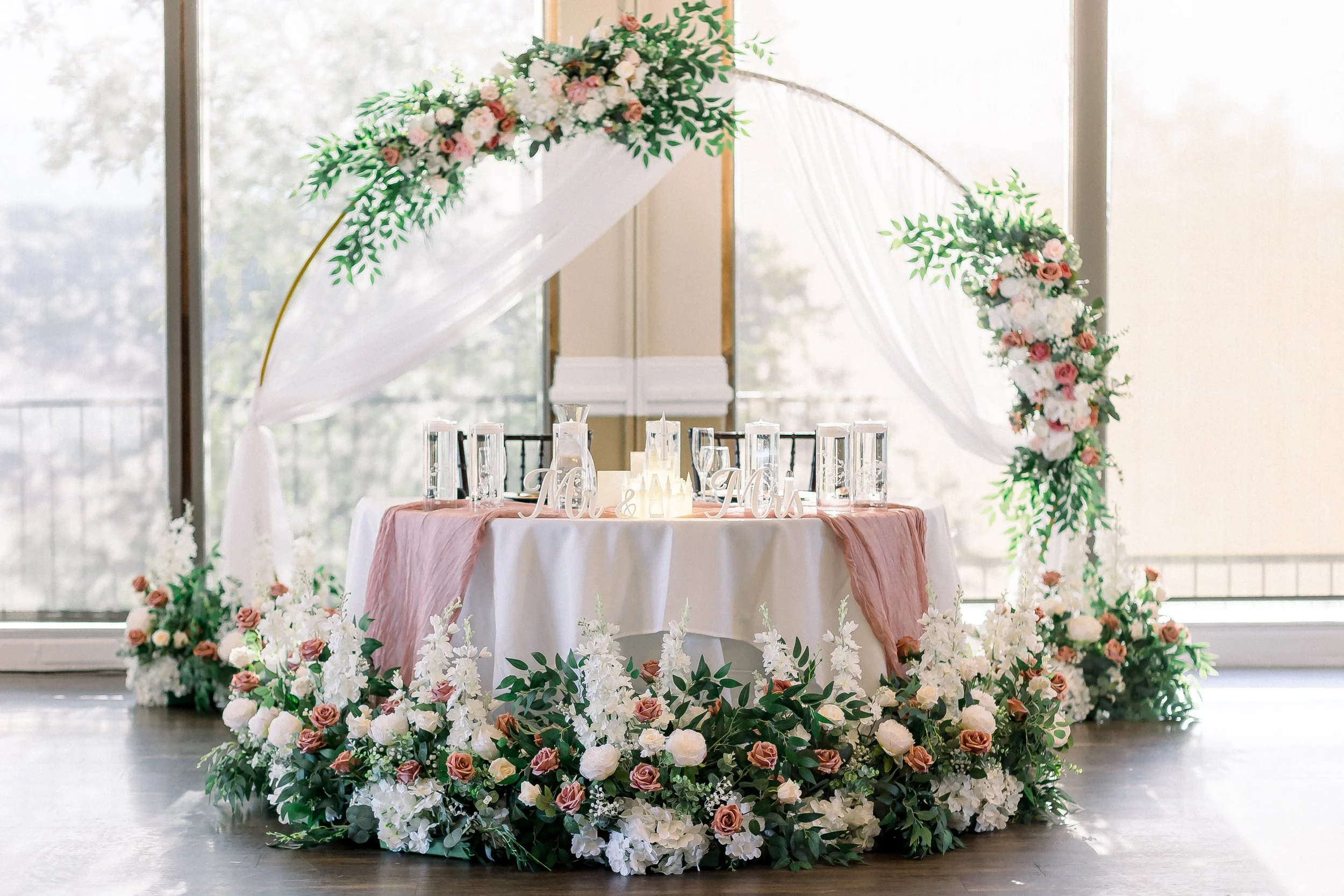 A decorated wedding or event table with floral arrangements, candles, and a sign that says 'Mr. & Mrs.' in a bright room with large windows and natural light.