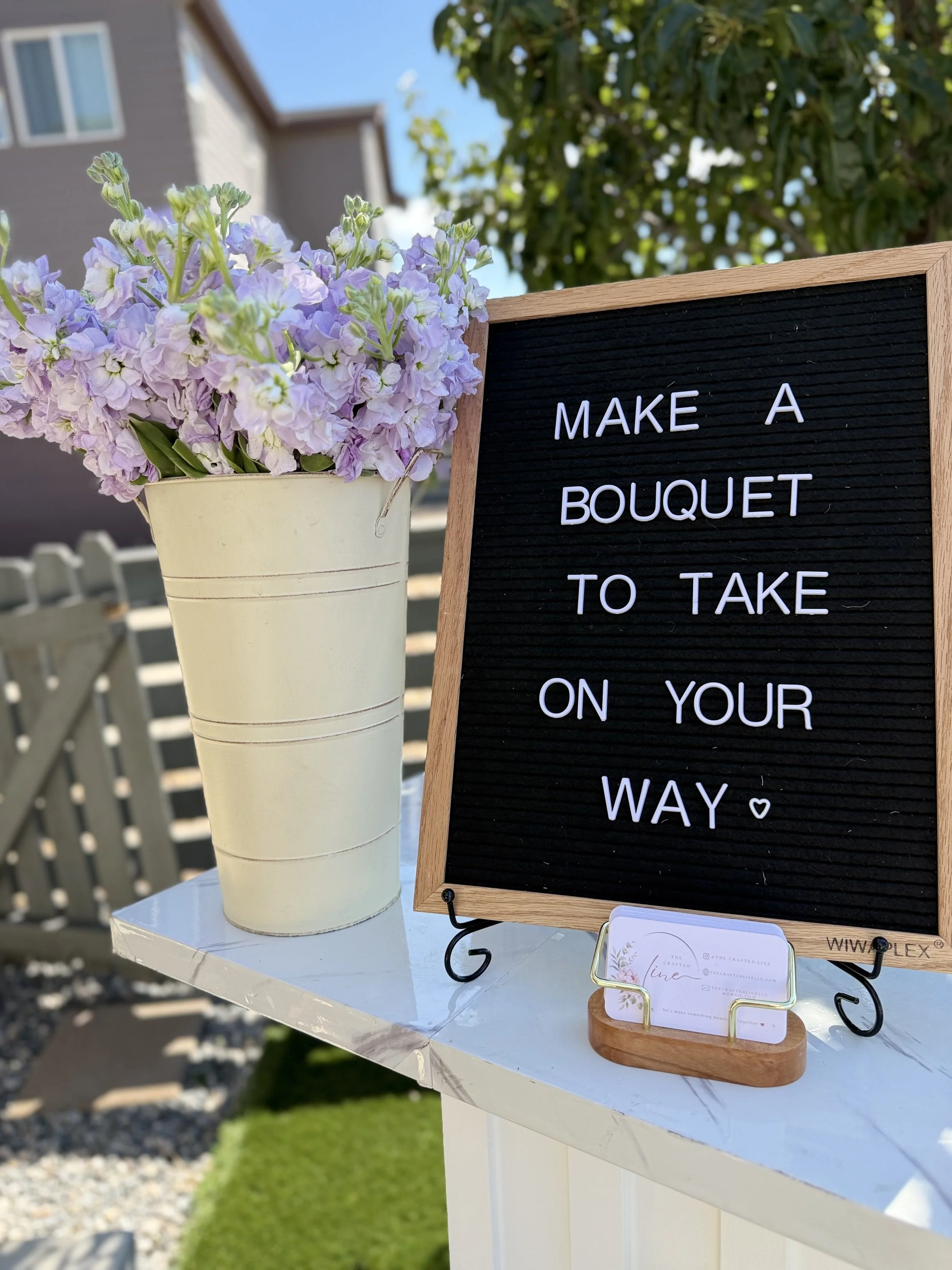 A curated bouquet station featuring white containers filled with pink roses, lavender stems, and seasonal florals. Includes a letter board sign inviting guests to “Make a Bouquet to Take on Your Way.”