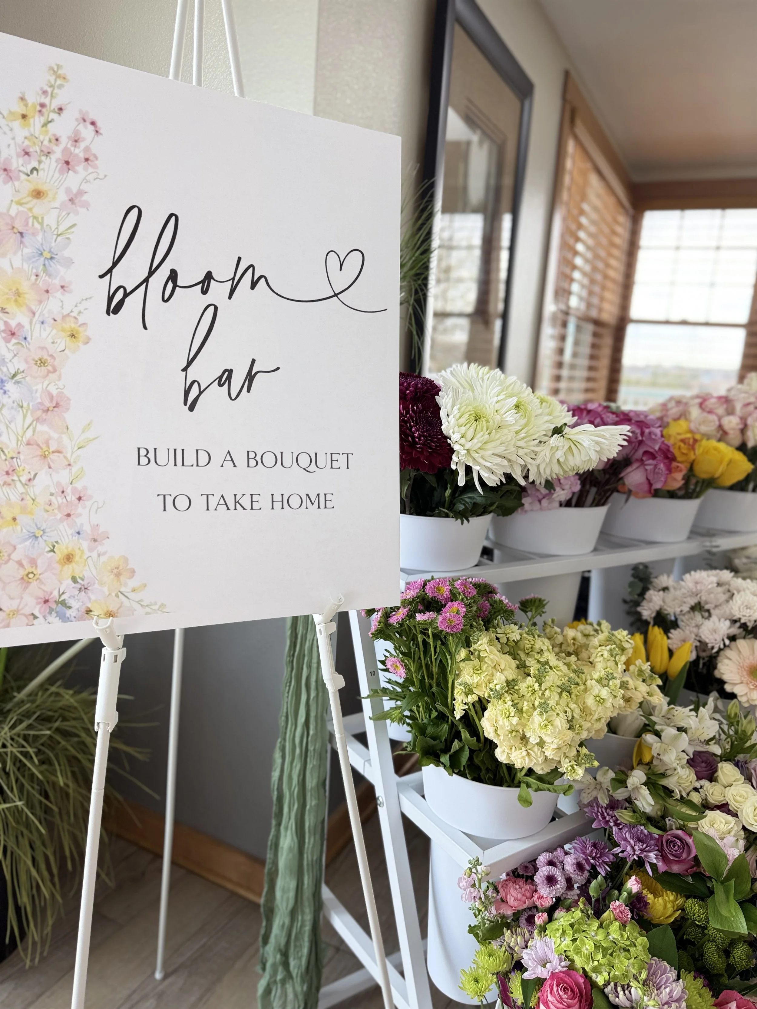 A display of colorful flowers in white containers at a floral shop, with a sign that reads 'Bloom & Bar: Build a bouquet to take home'.