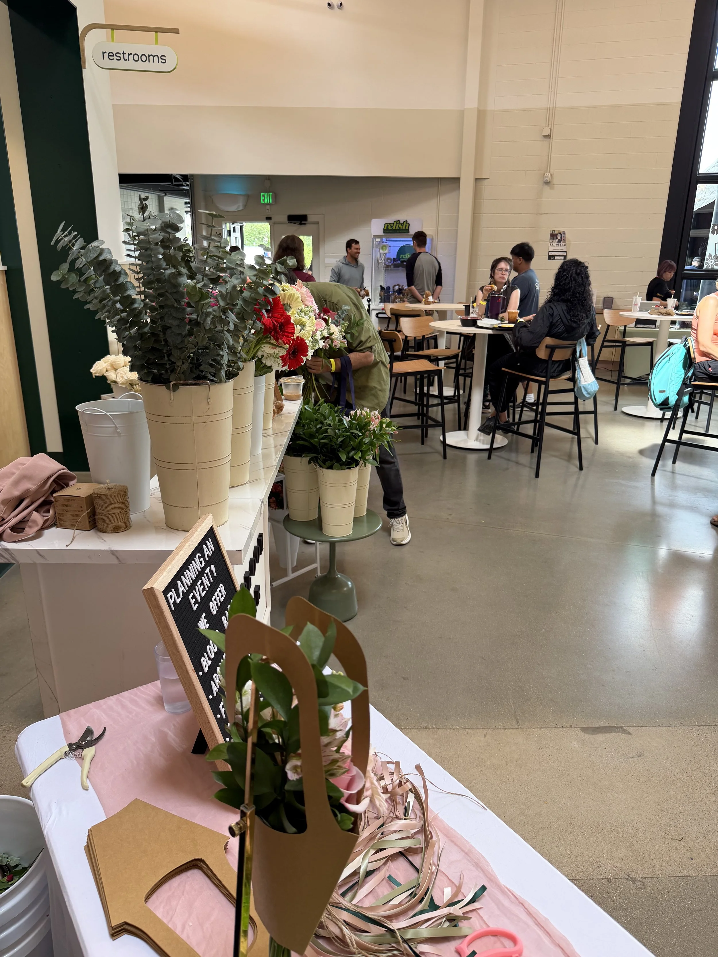 Display of floral arrangements and crafting supplies on a table in a community space, with people socializing and a sign indicating restrooms in the background.