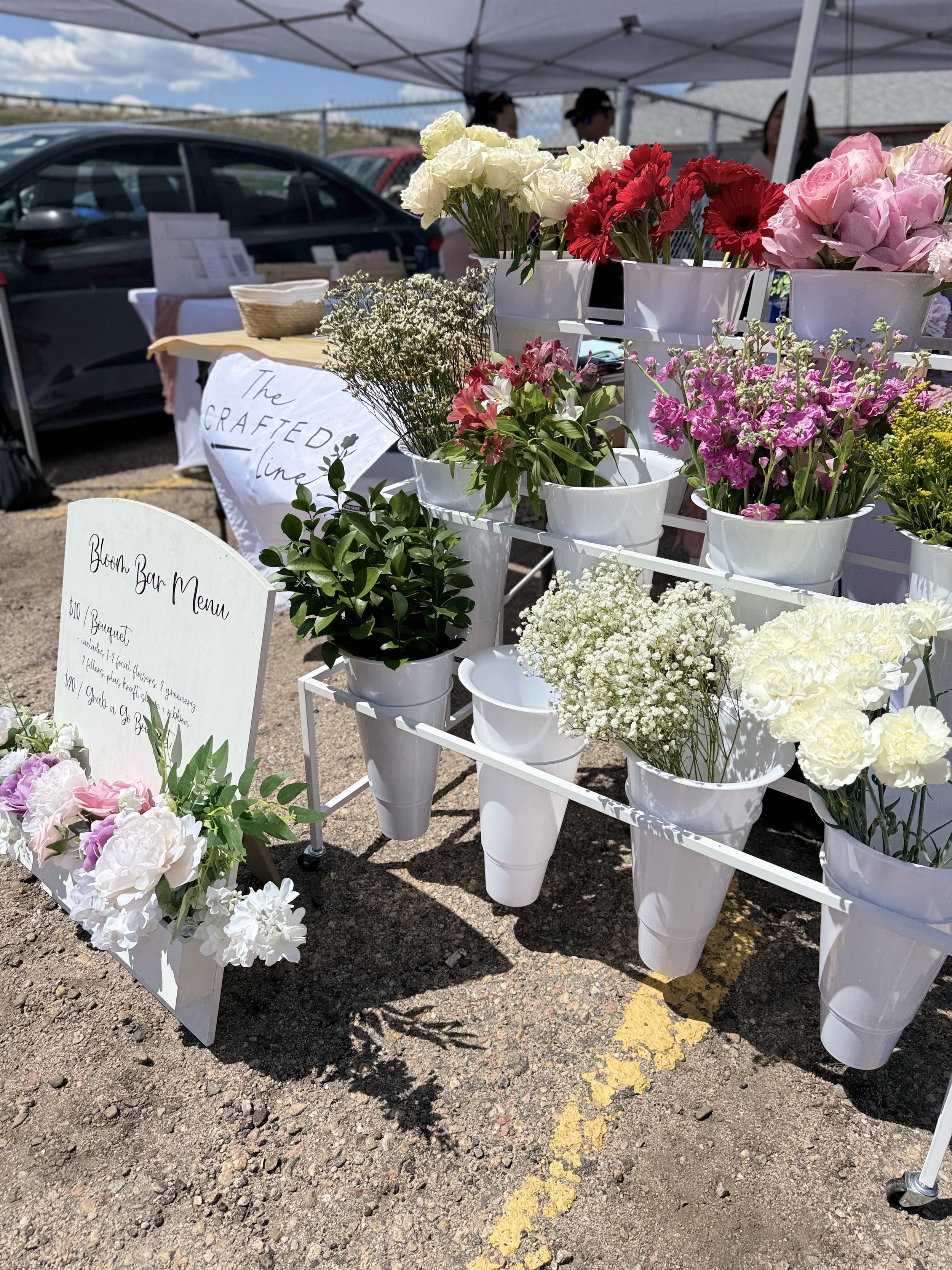 A display of various colorful flowers at an outdoor market stall, with a signboard and a few people in the background.