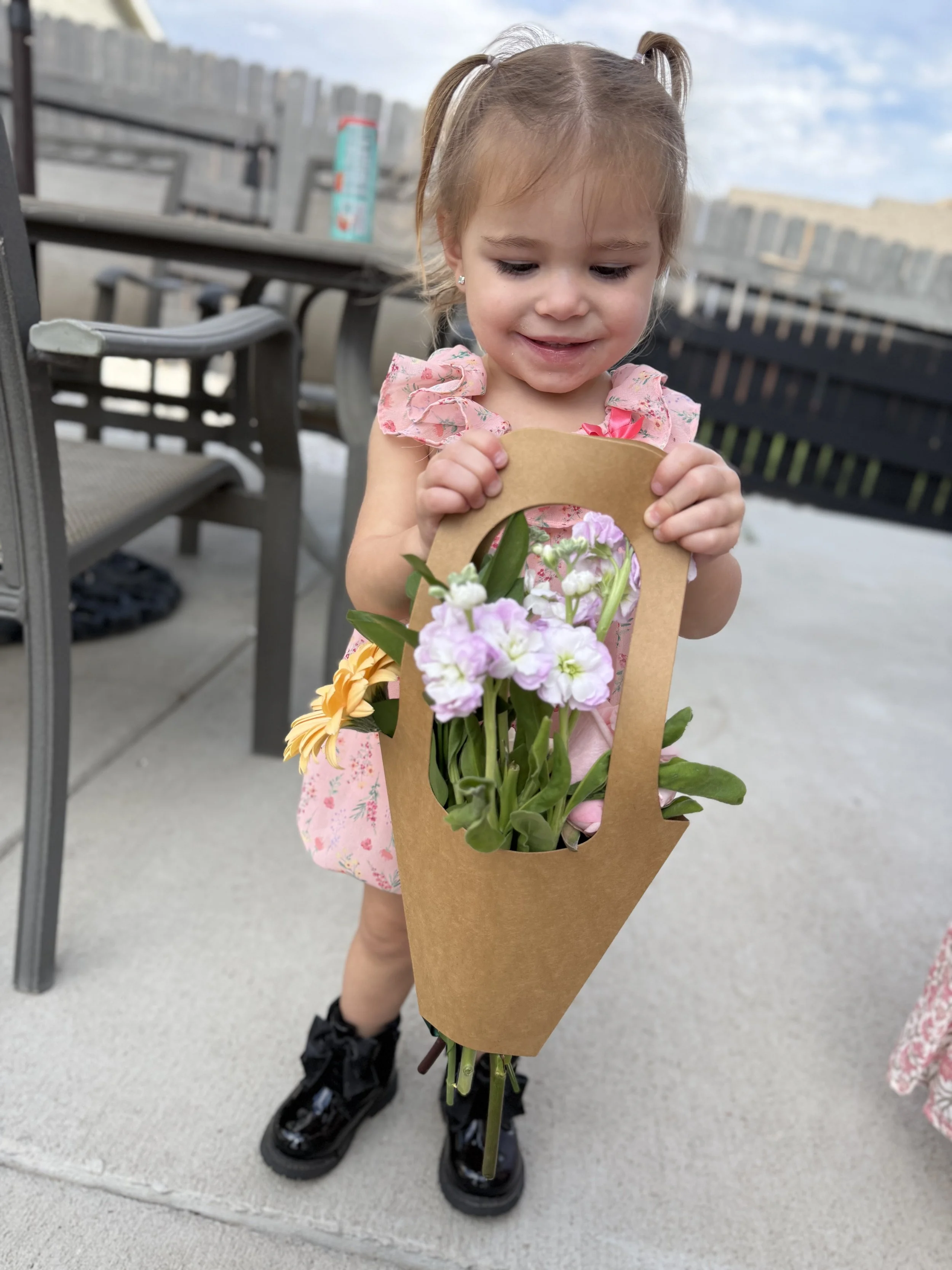 A young girl in a pink floral dress and black shoes holding a brown paper bag with purple, white, and yellow flowers inside.