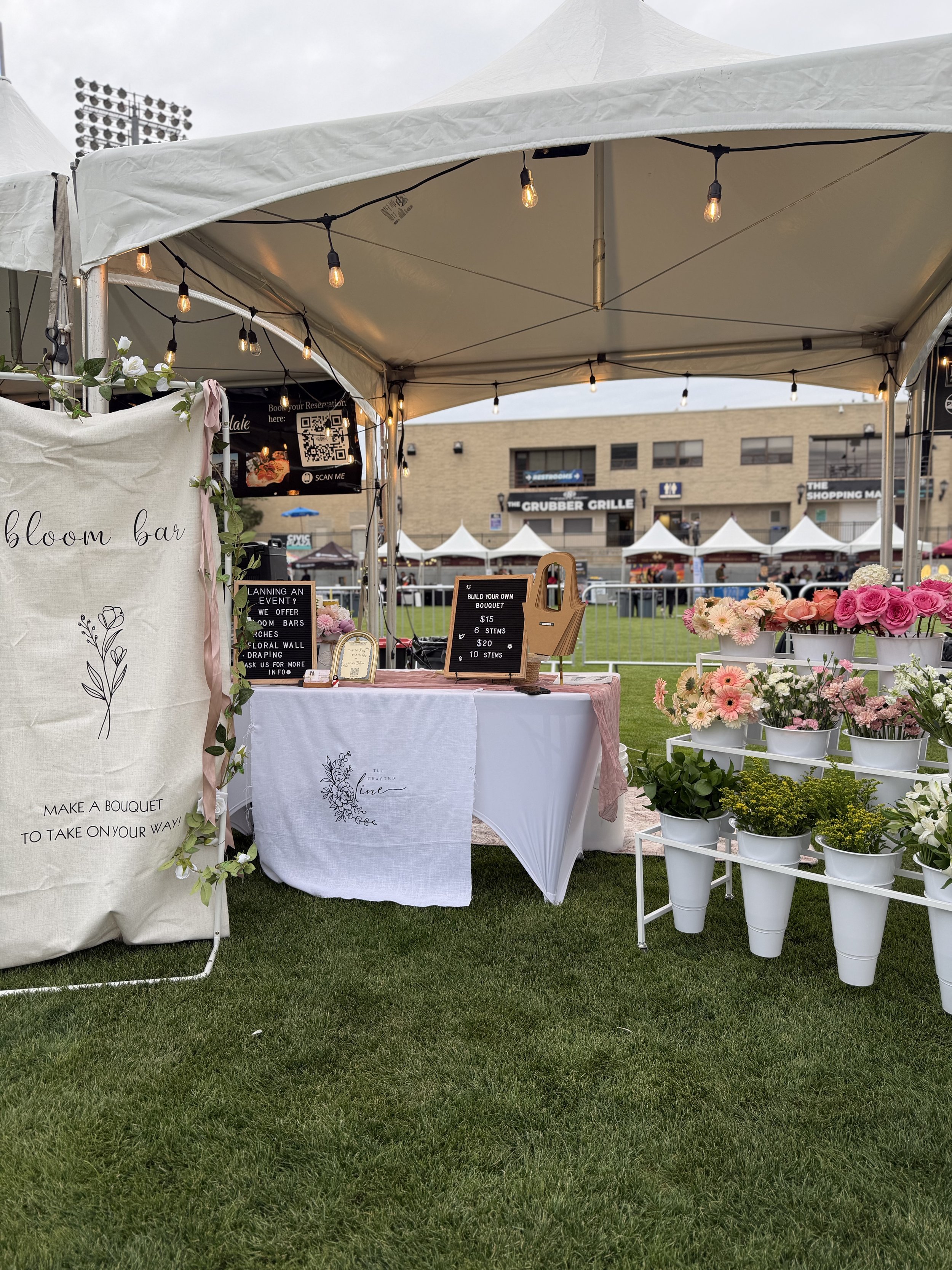 A lively outdoor market display with tables draped in white cloths and buckets overflowing with colorful flowers. The tented setup feels warm and welcoming, showcasing a vibrant selection ready for guests to build their own bouquets.