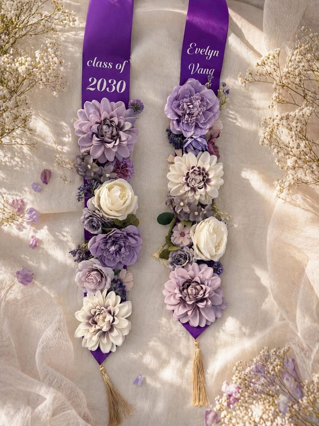 Graduation sashes decorated with purple, white, and lavender flowers, with text indicating the class of 2030 and the name Evelyn Vang, laid on a soft fabric with baby's breath flowers around.